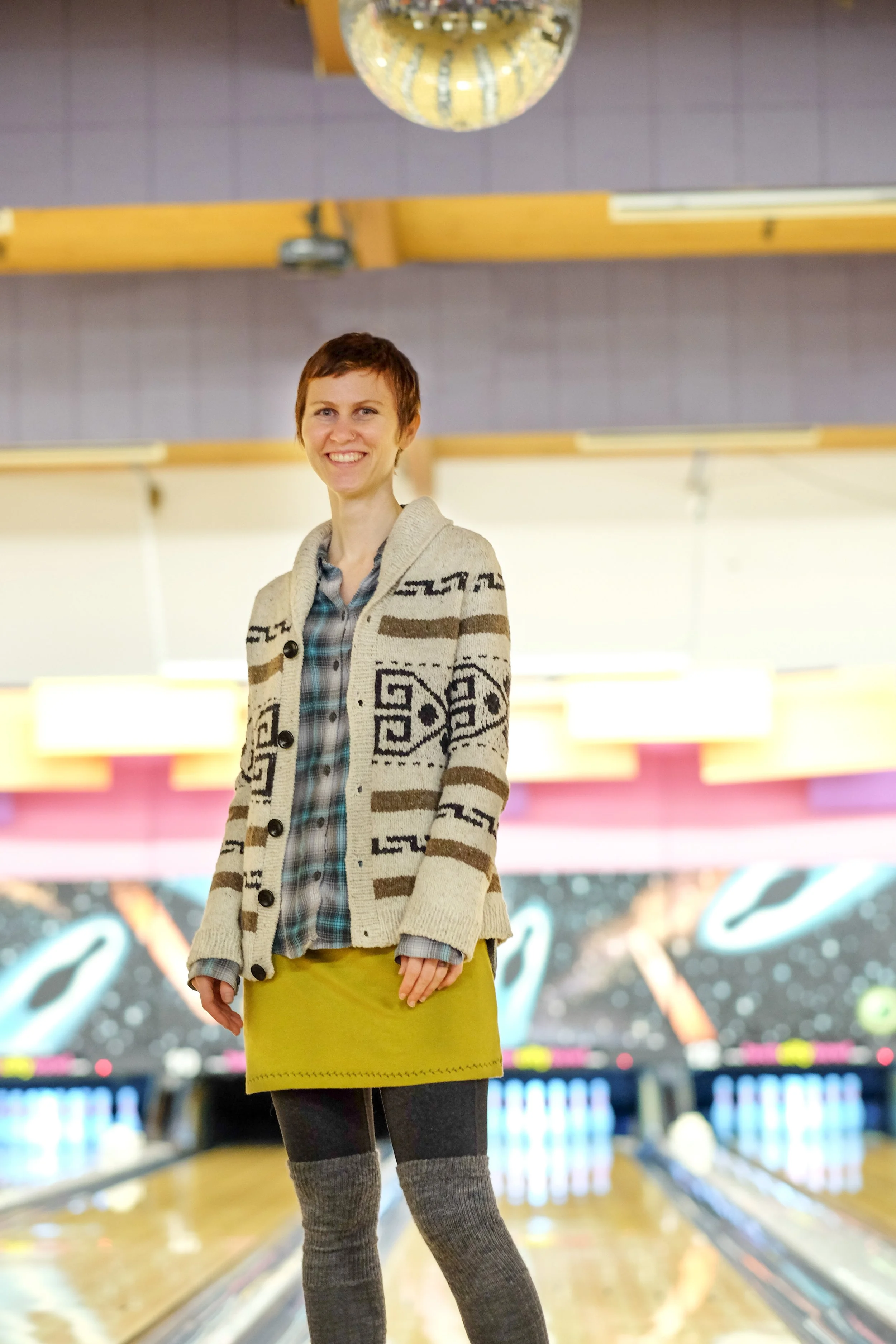 Andrea with a very short pixie haircut is standing in a bright bowling alley wearing her Knitter’s Dude sweater, a mustard-coloured miniskirt, and over-the-knee socks.