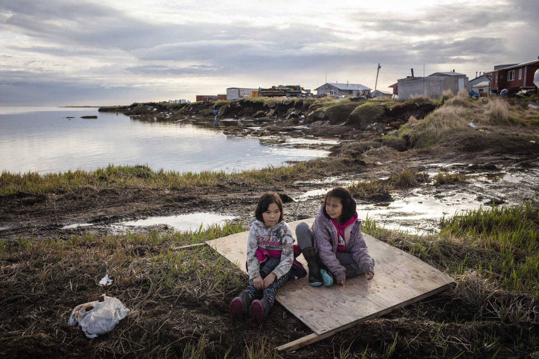  Clarissa and Aaliyah sit on top of a plywood board used to walk safely on sinking mud in Newtok, Alaska. May 28th, 2019. Behind them are crumbling permafrost cliffs that have become too dangerous for nearby homes. At the time, multiple unsafe homes 
