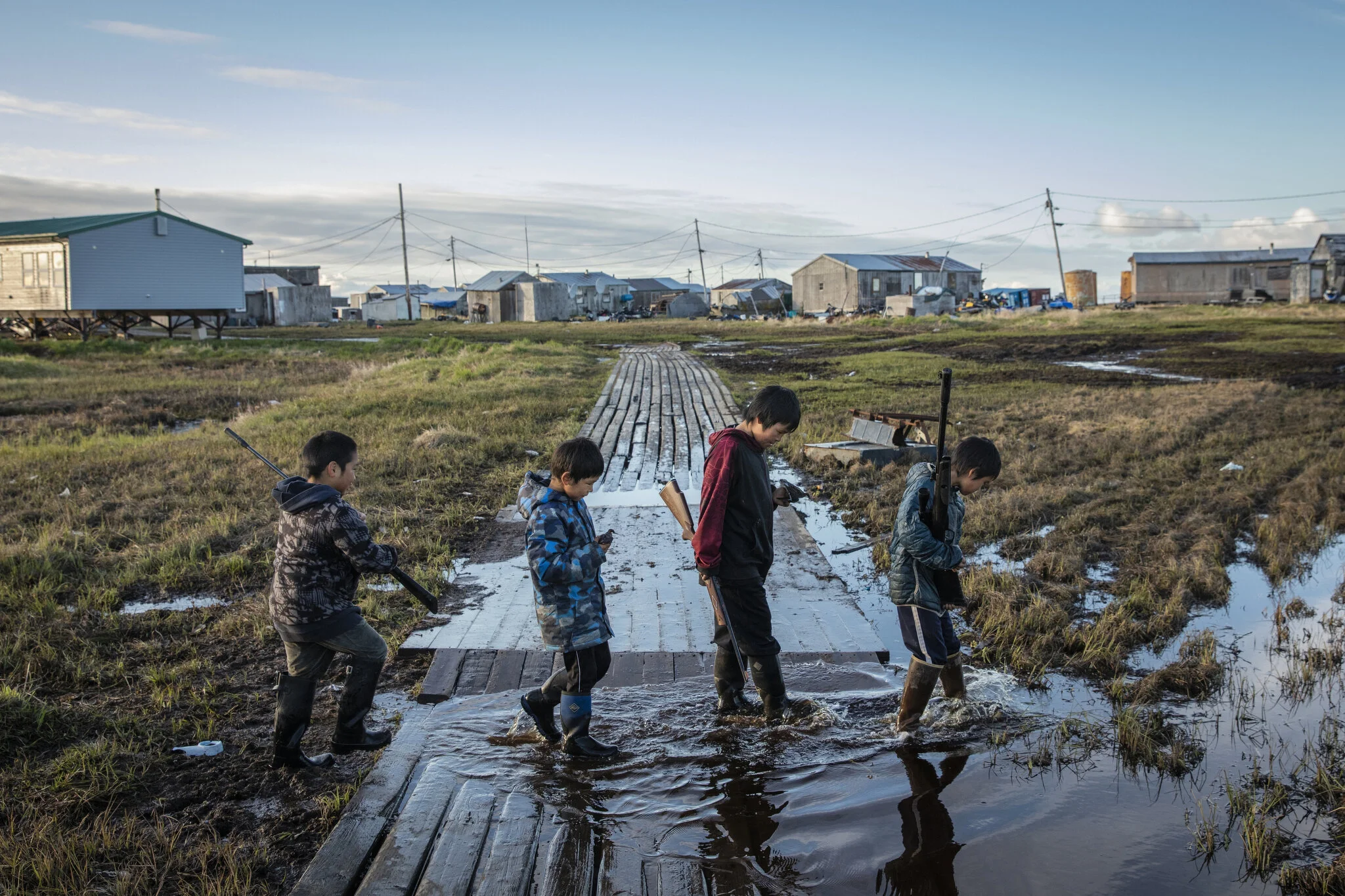 On a summer bird hunt, Kenyon Kassaiuli, Jonah Andy, Larry Charles, and Reese John cross a flooded walkway in Newtok, Alaska. May 27th, 2019. The Yupik village of Newtok, Alaska, population 380, is sinking as the permafrost beneath it thaws. Erosion