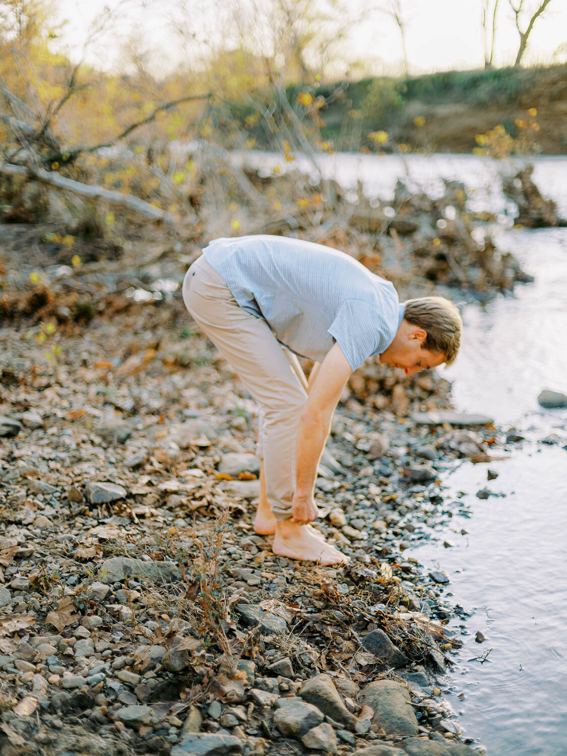 man-rolling-up-slacks-to-wade-in-rivanna-river-during-golden-hour-at-his-engagement-session-in-charlottesville-virginia.jpg