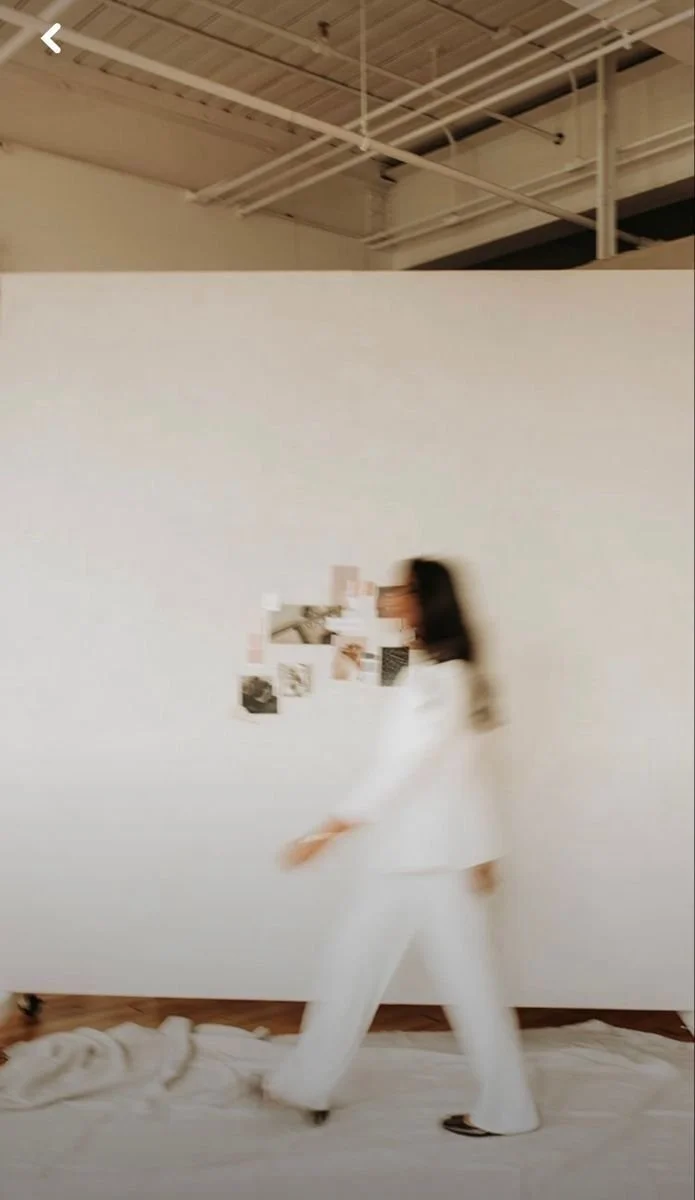 A person with long dark hair walking past a large white wall with small photographs and images attached to it, in a room with an exposed ceiling and white pipes.