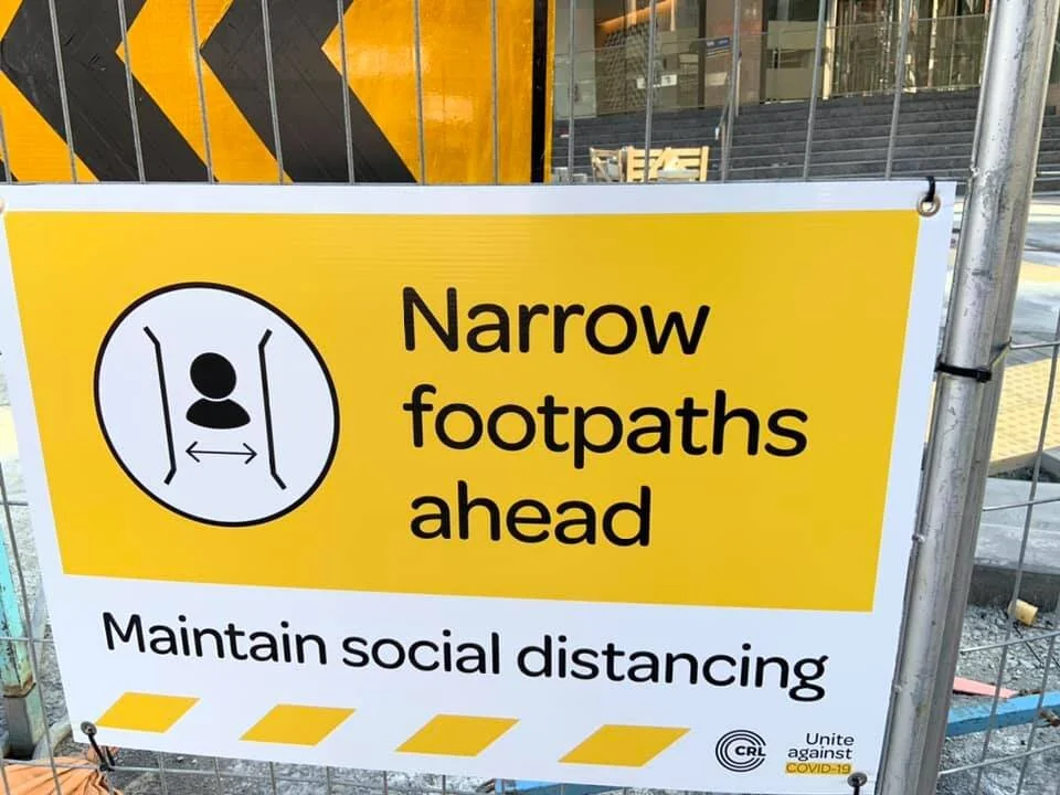 Picture of yellow and black COVID-19 sign on fence advising of narrow footpaths ahead for social distancing purposes