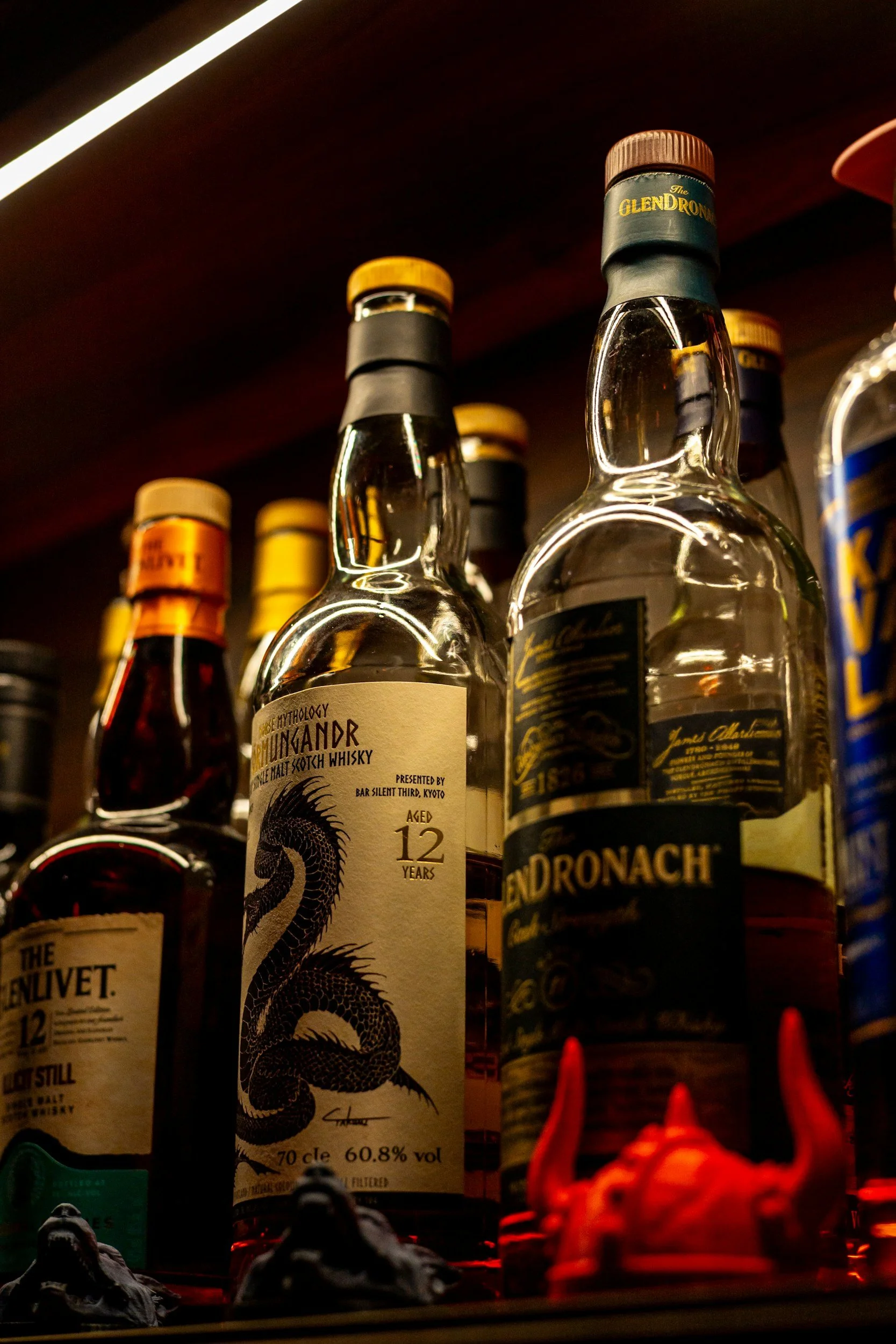 Several bottles of alcohol on a shelf, including a Scottish whisky with a white label and a black dragon illustration, and other bottles with black and gold labels, some with orange and red cap tops, illuminated in warm lighting.