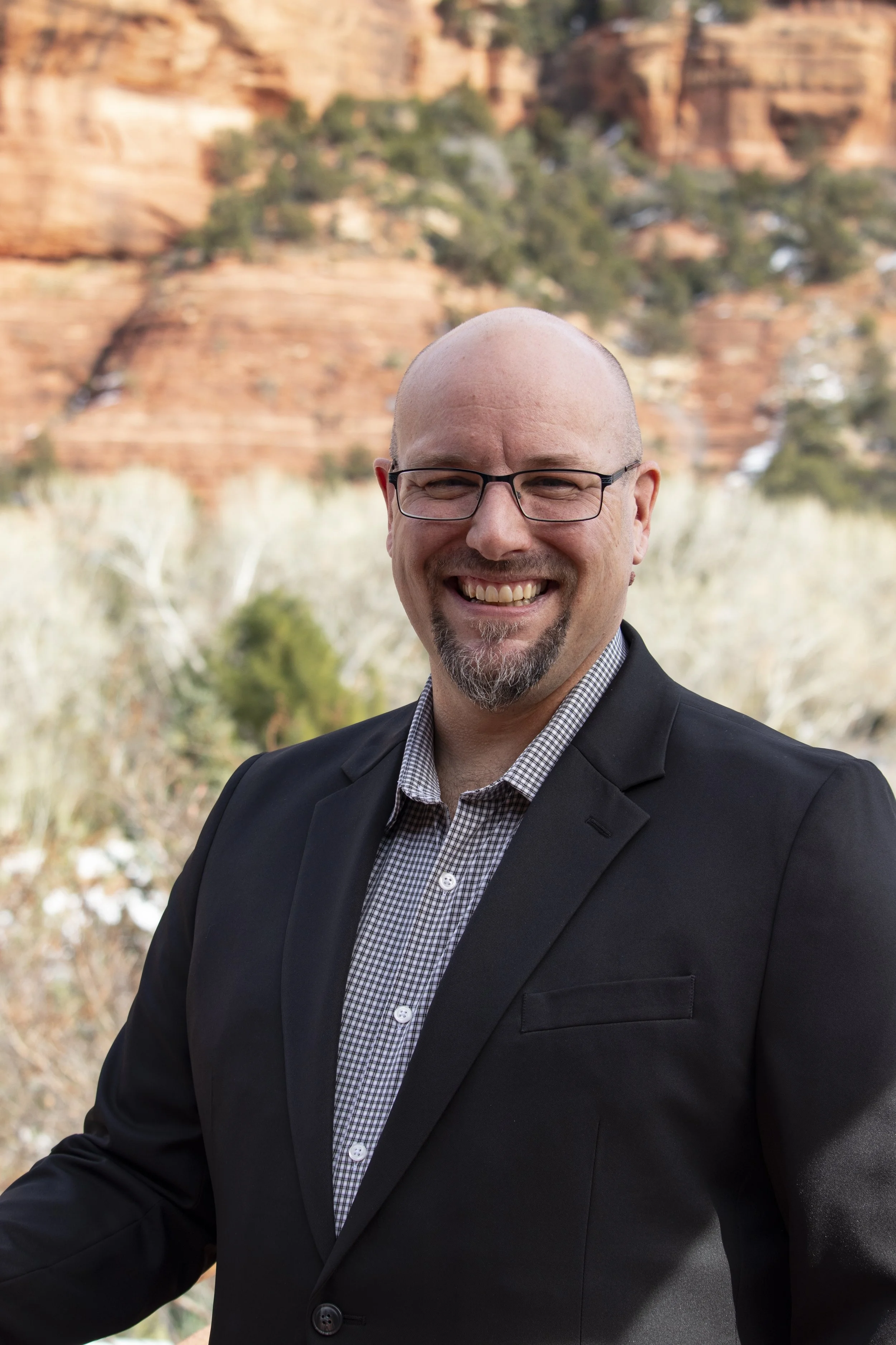 A smiling man in a plaid shirt and a black blazer standing outdoors with a red rock canyon and trees in the background.