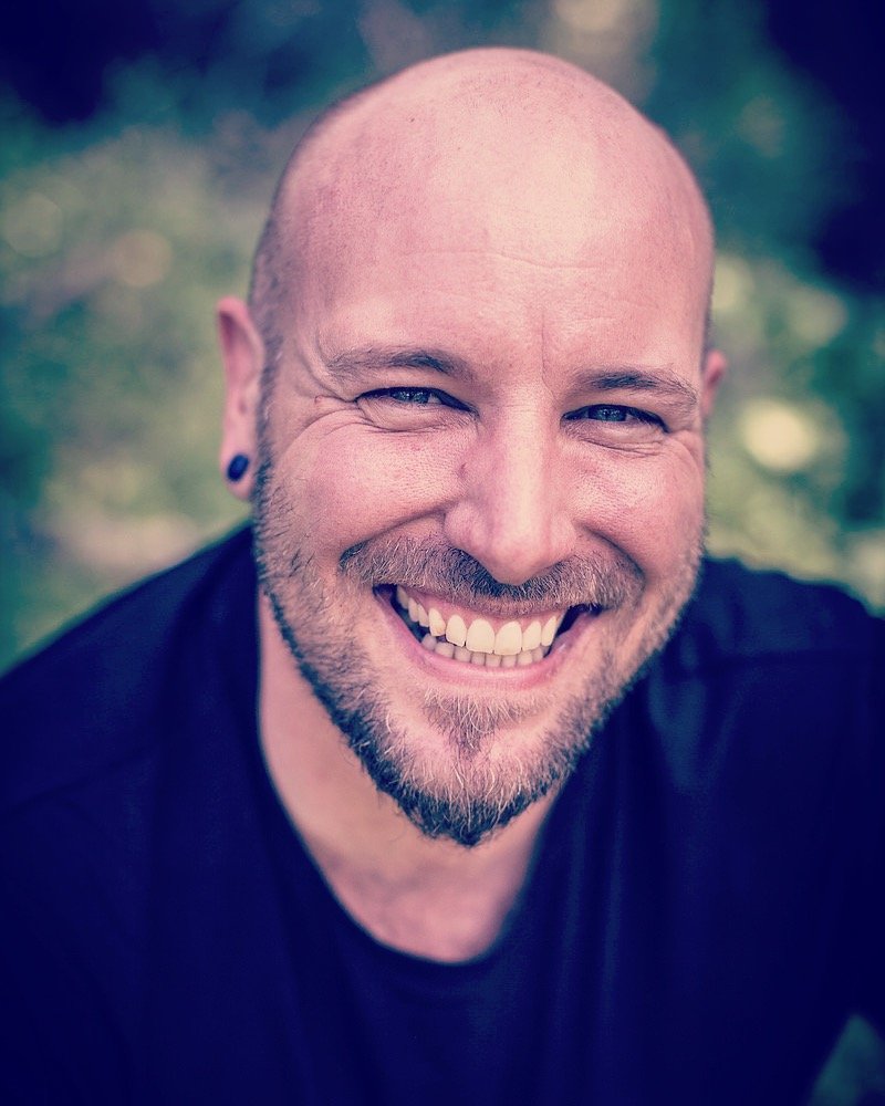 A smiling man with a bald head and a beard, wearing a black shirt, outdoors with blurred trees in the background.