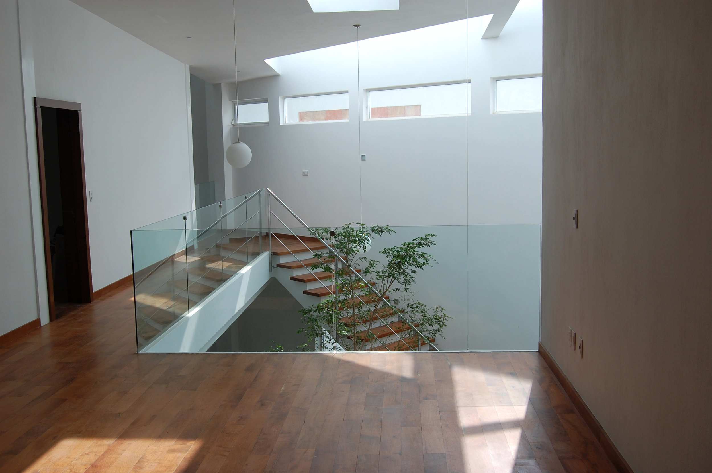 Is a residential house located in the Valle Real neighborhood, in Guadalajara, Mexico. The project starts from the idea of achieving the maximum transparency and visual relief towards the dense wooded area that adjoins the back of the property. Likewise, it is intended to preserve some privacy and discretion in front, towards the street. This was achieved not only by the distribution and orientation of its main spaces towards the transparency of the back façade but also by organizing the interior around a double height space that embraces the main staircase and an indoor tree who's presence embellishes the space echoes the importance of the neighboring trees. Thus the circulations of the house both upstairs and downstairs open and breath into the space of the inner tree while its main spaces are directed and flow towards the wooded area on the back.