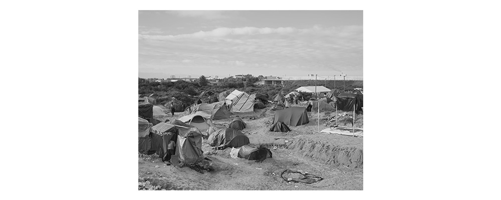 Image I – The Jungle The largest illegal refugee camp in Western Europe is just a few hours away from us in Calais. It is a gathering point for refugees who risk their lives to get through the tunnel or by ferry to England. At the time of this photograph, an estimated 3,500 were „residents“ in the „Jungle“. Police and fences with razor wire make it virtually impossible to reach a ferry or the Euro Tunnel. Nevertheless, every evening, refugees leave their tents and risk their lives to somehow continue their journey. The camp itself is a legal vacuum. We could only enter it safely in the morning. The closer the time of the evening departure, the more aggressive and unpredictable the mood gets. The frustration of failure then often discharges at night, culminating in violent riots like in May 2016, with many injured. Despite several attempts to evacuate the camp, about 4,000–5,000 refugees still live in the dunes of Calais today.