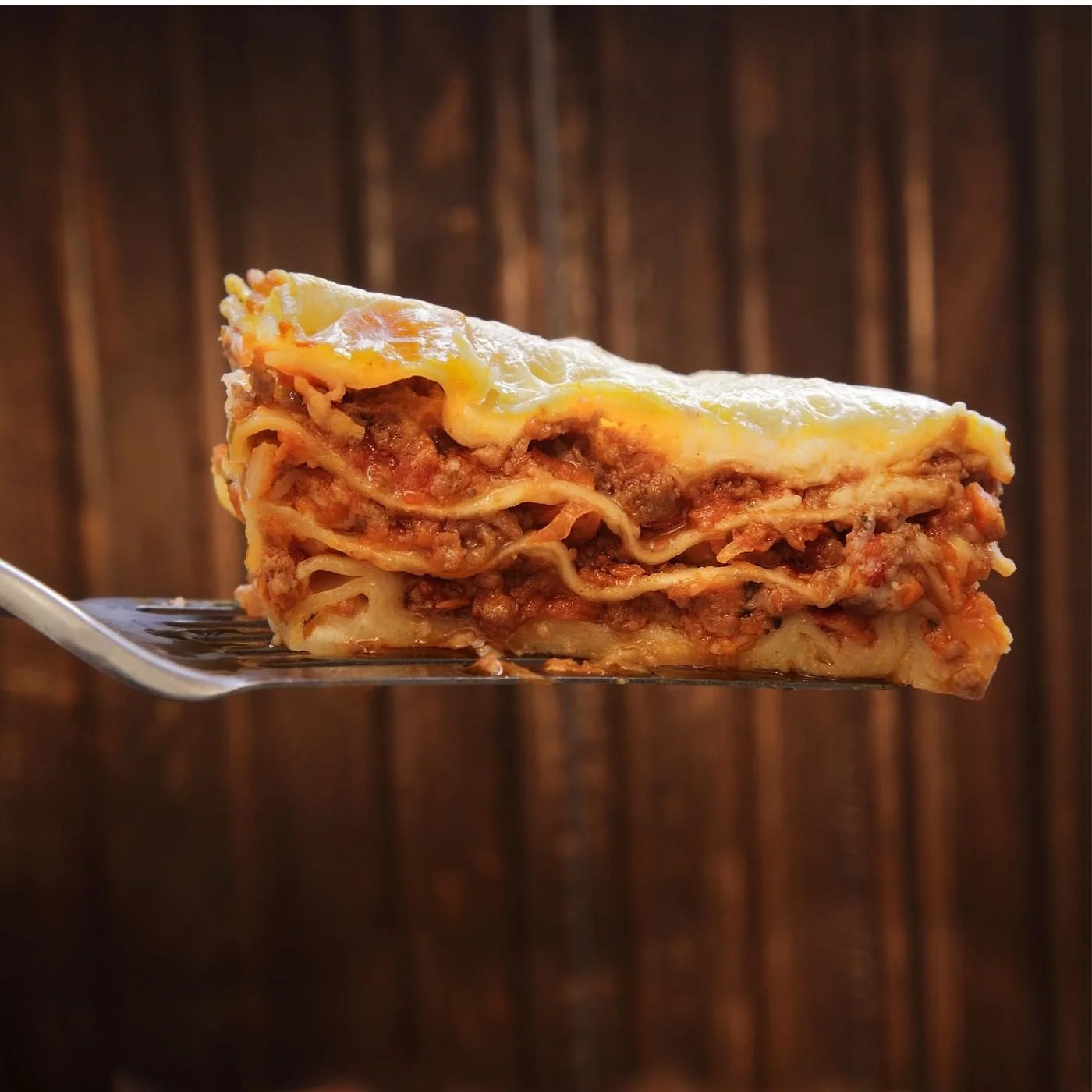 a lasagna slice on a metal spatula held in front of a wooden background