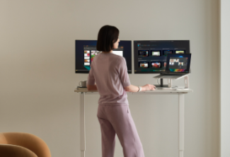 Woman with brown hair at standing desk with two monitors and wearing a pink suit. Beige background.