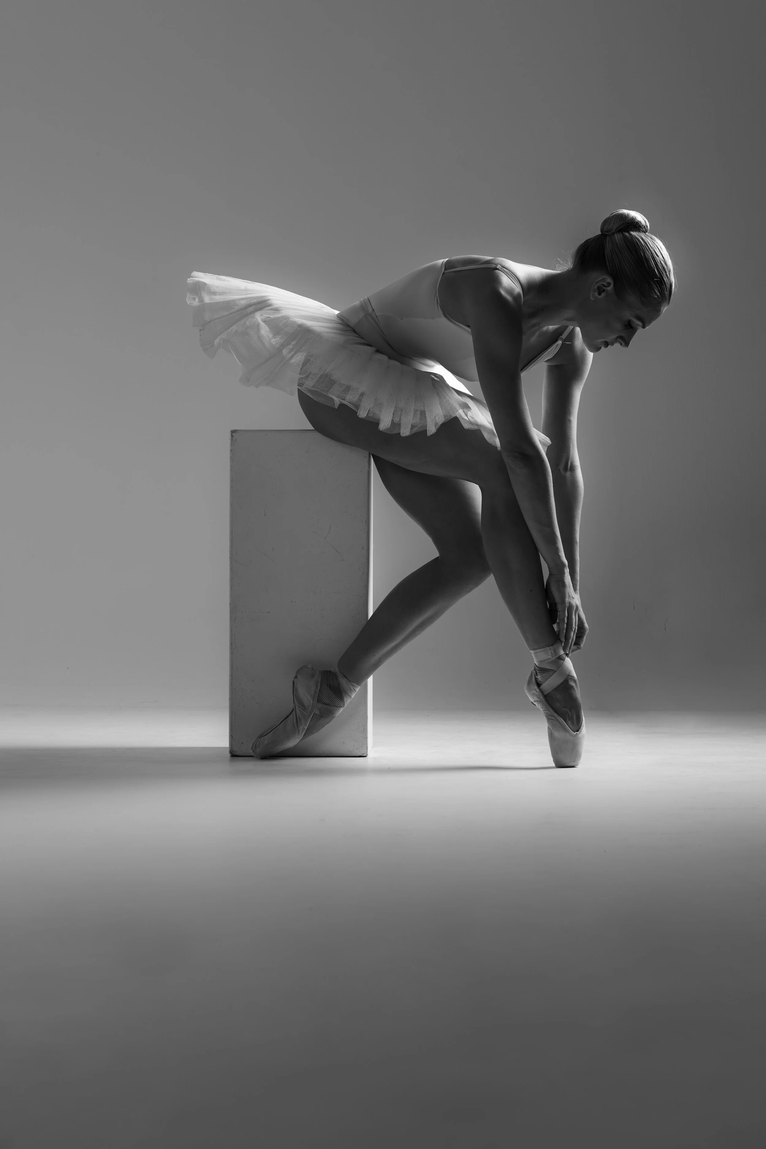 A ballerina in a tutu stretching on a ballet block in a studio, captured in black and white.