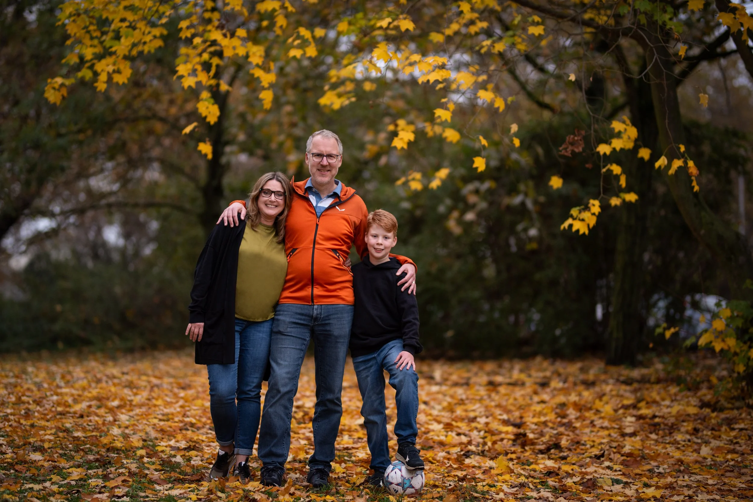 Family of three standing together outdoors in autumn, surrounded by fallen leaves and trees with yellow leaves, smiling at the camera.