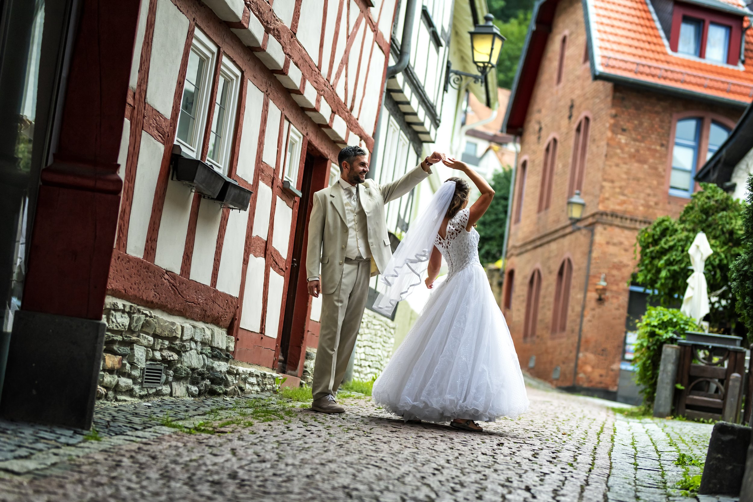 A newlywed couple dances on a cobblestone street in front of historic half-timbered and brick buildings, with trees and street lamps in the background.