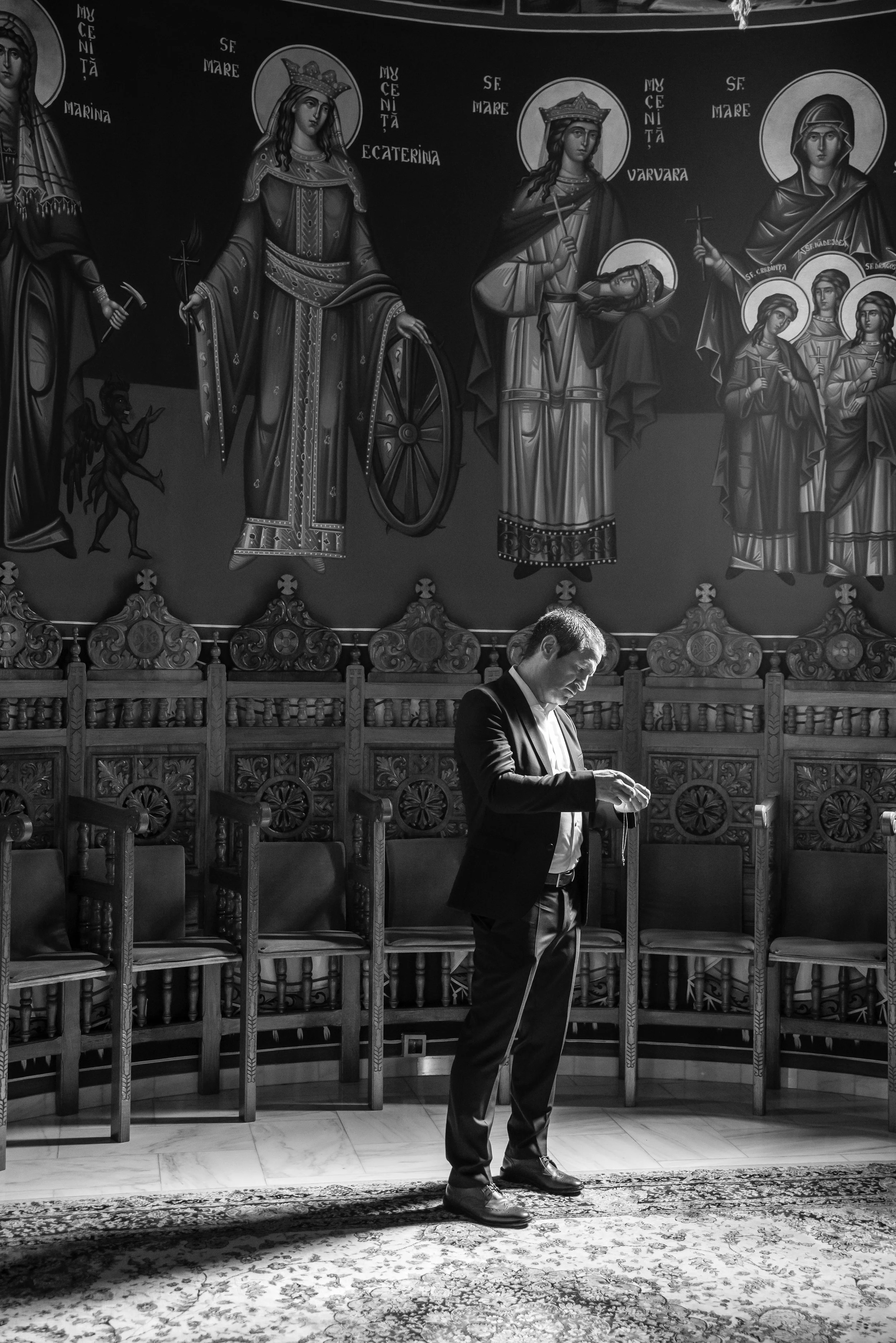 A man in formal attire stands in a room with ornate wooden chairs, illuminated by a beam of light, with religious iconography on the wall behind him. UsArtFoto Wedding photography
