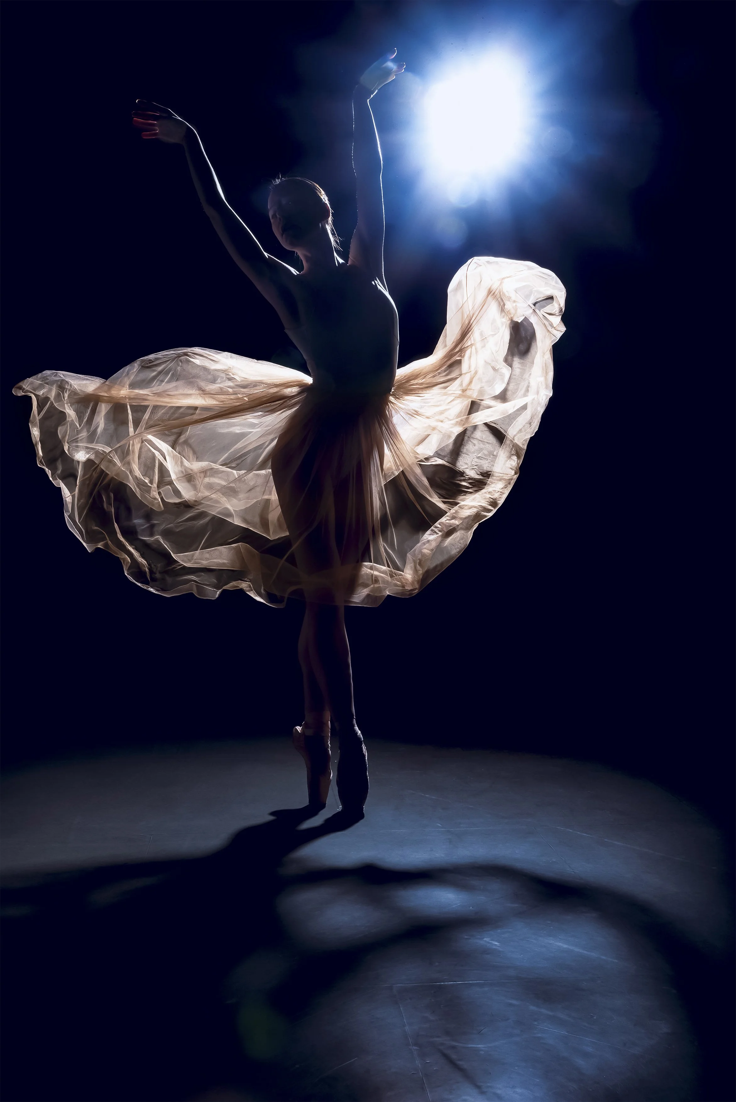 A ballet dancer in a beige tutu performing on stage with backlighting against a dark background.