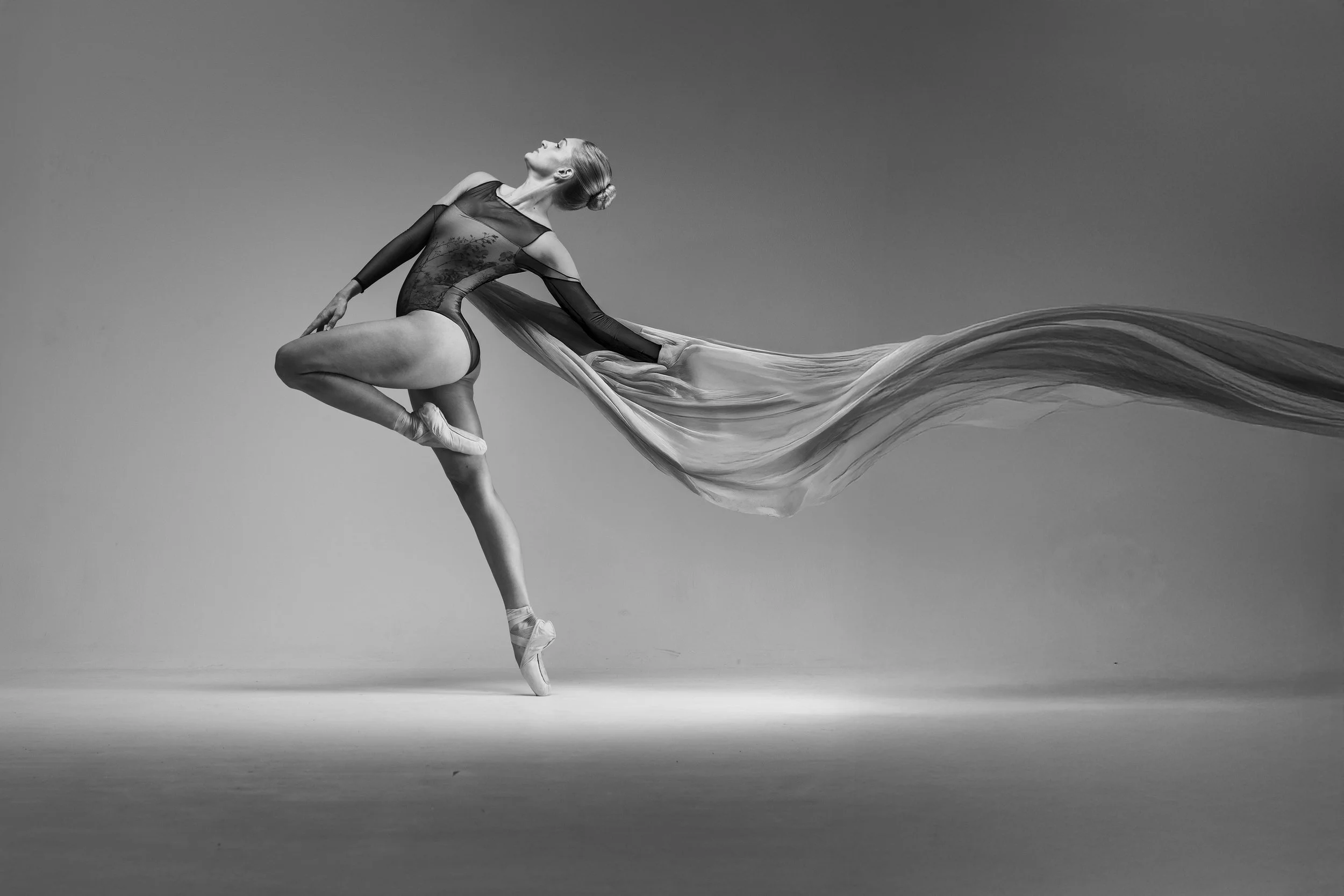 A ballerina in mid-pose on pointed toes, wearing a leotard with a flowing fabric extending from her back, in a black and white studio shot.