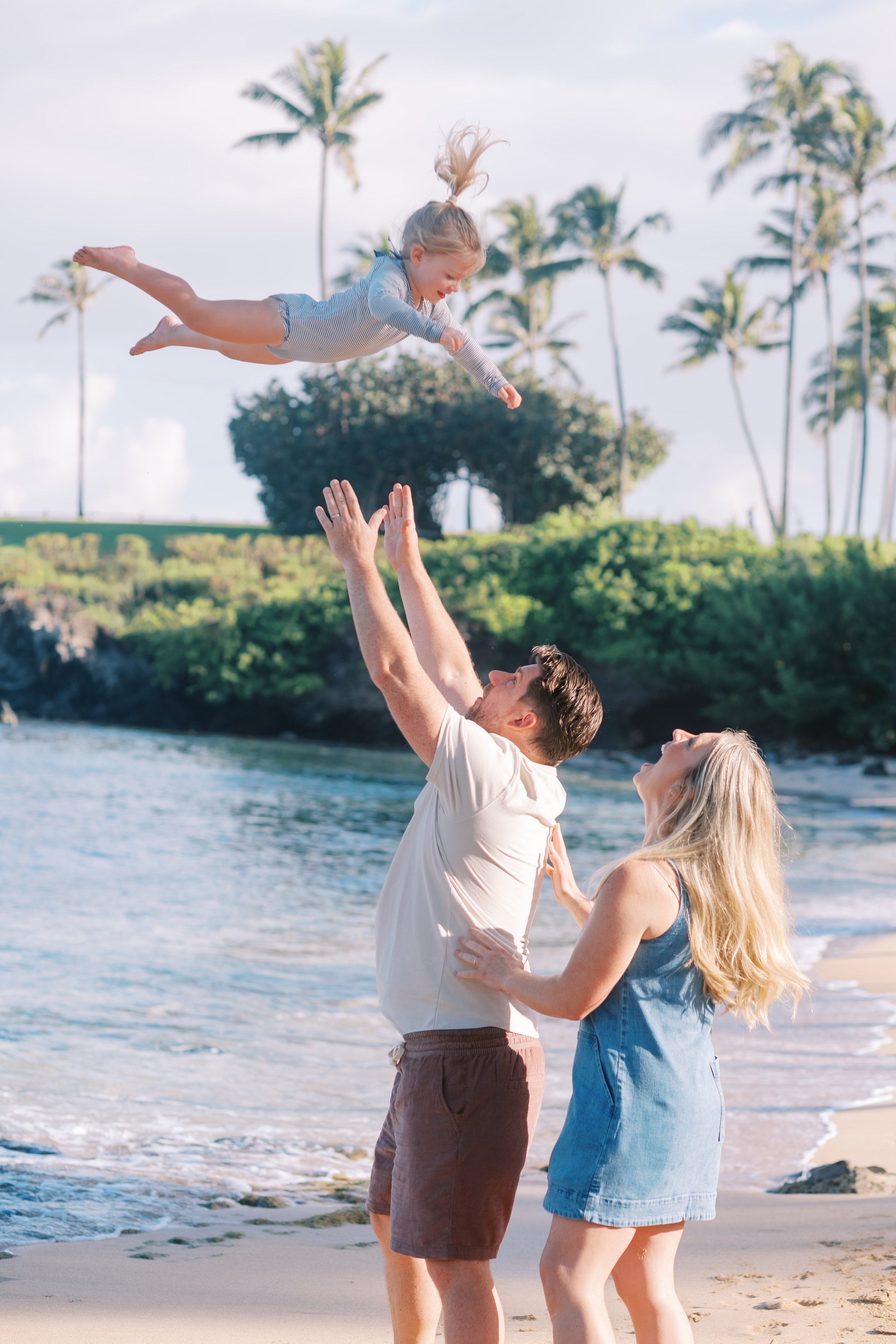 A family photoshoot at Kapalua Bay with a little girl being tossed in the air by her father while laughing.