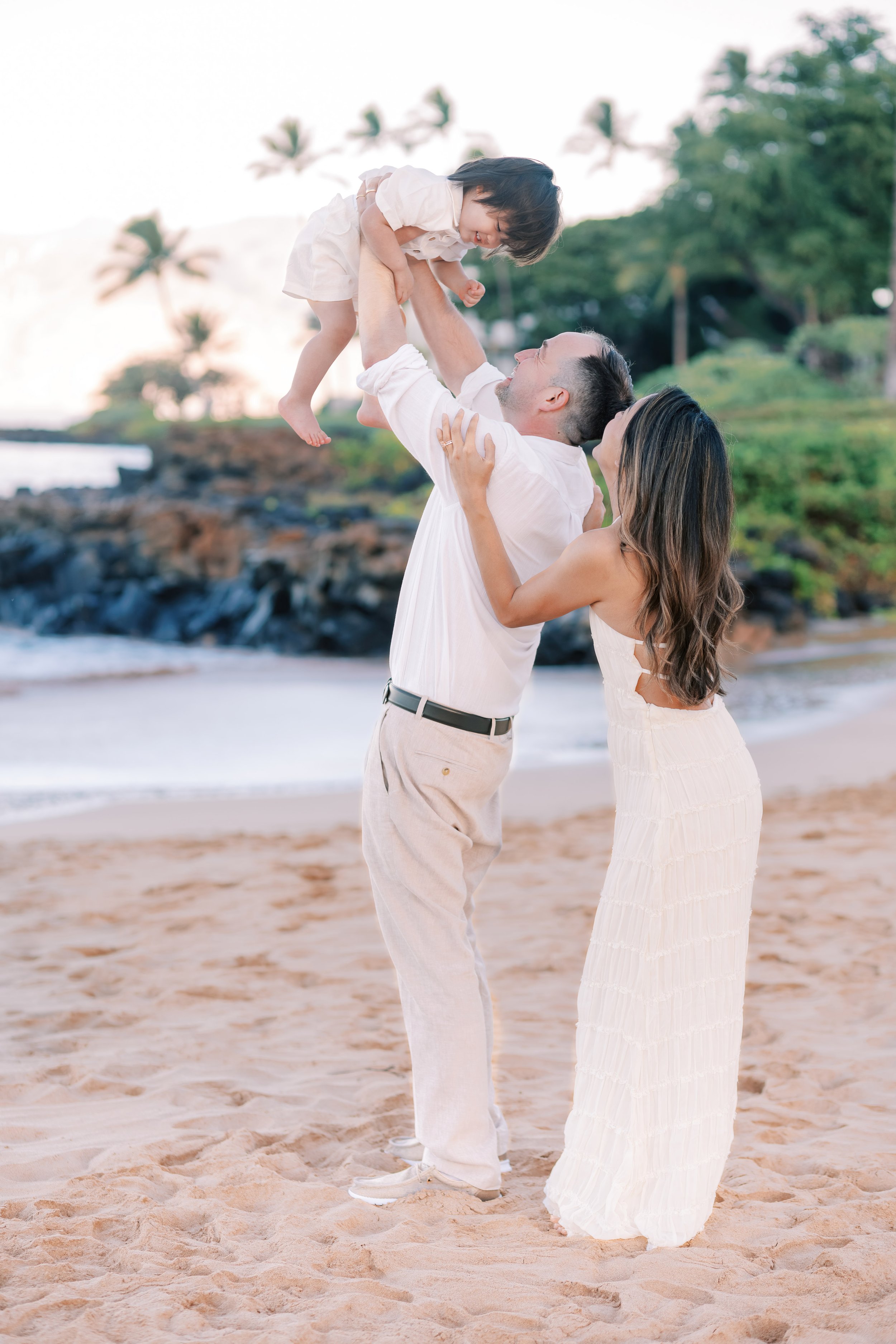 A father holding up his toddler who is giggling during a family photoshoot on Wailea Beach in front of the Grand Wailea.