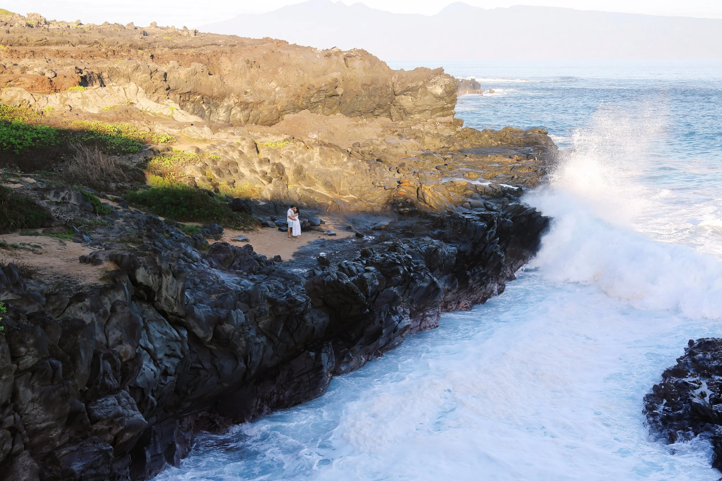 A couple kissing on the rocky cliffs at Ironwoods in Kapalua Maui with waves crashing below them during a morning photoshoot.