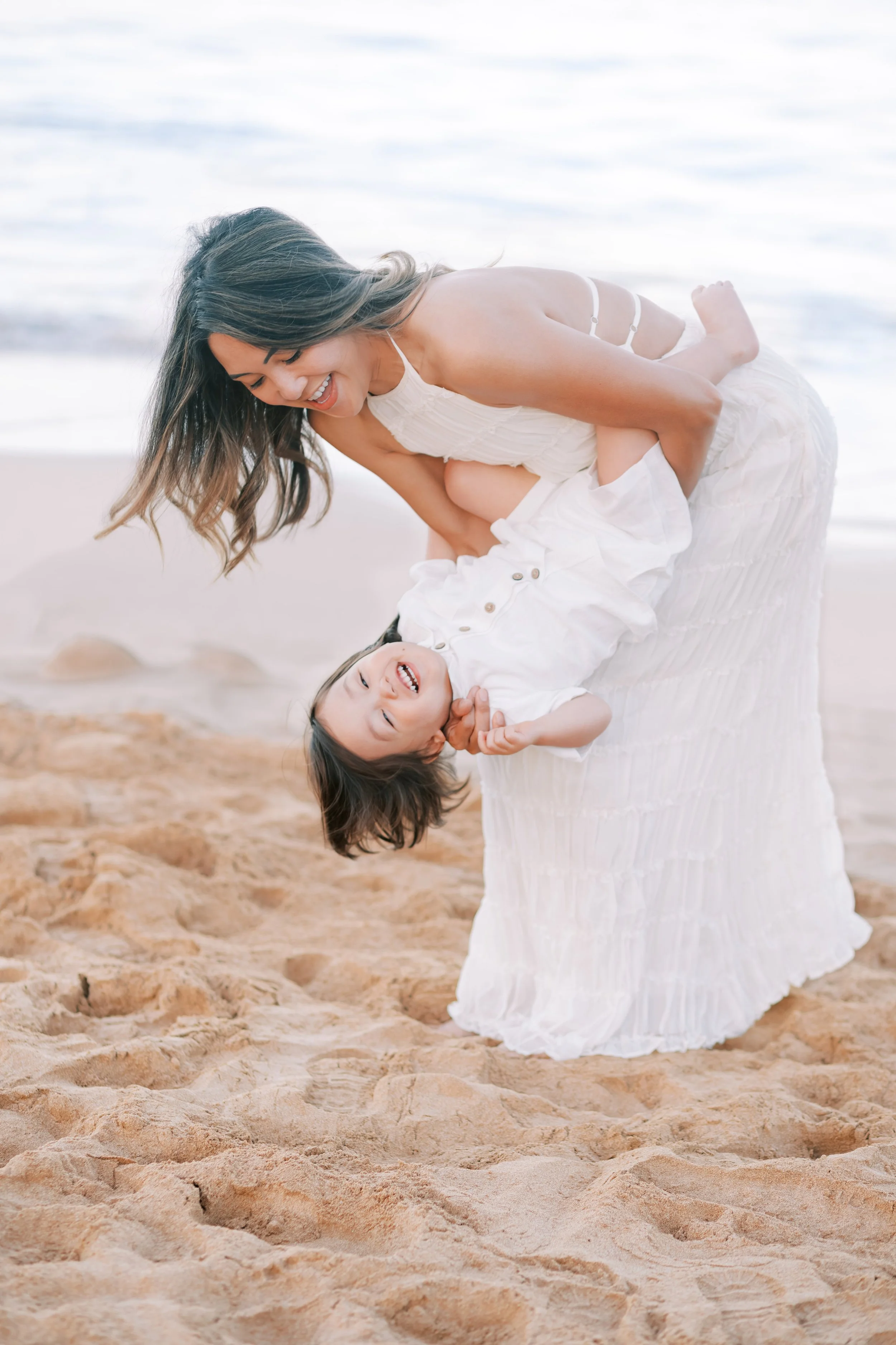 A mother dipping her giggling toddler during a sunrise photoshoot at Wailea Beach in front of the Grand Wailea in Maui.