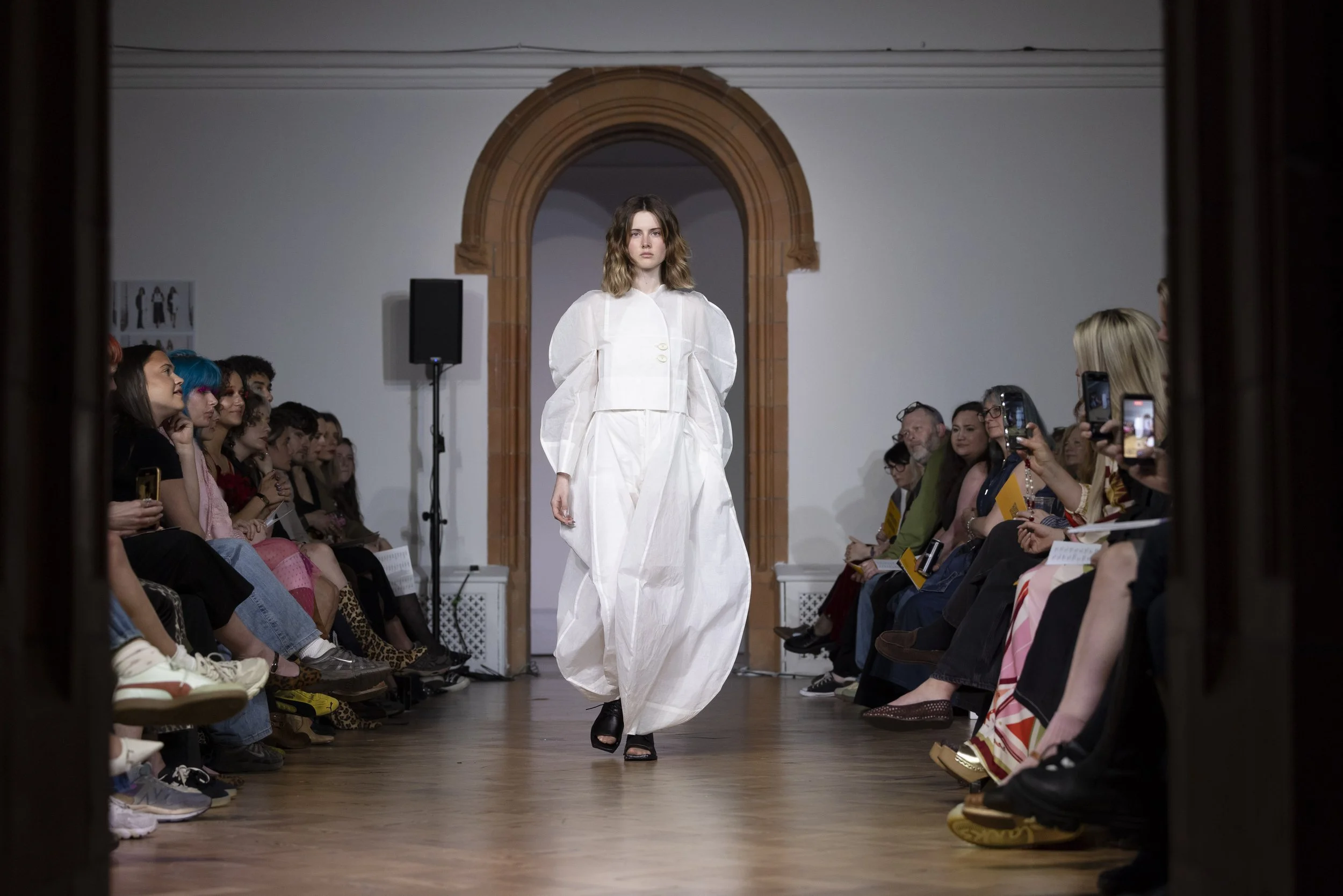 Model in a voluminous white dress walking down the centre of the catwalk at MMU fashion showcase, audience visible on both sides of the runway.
