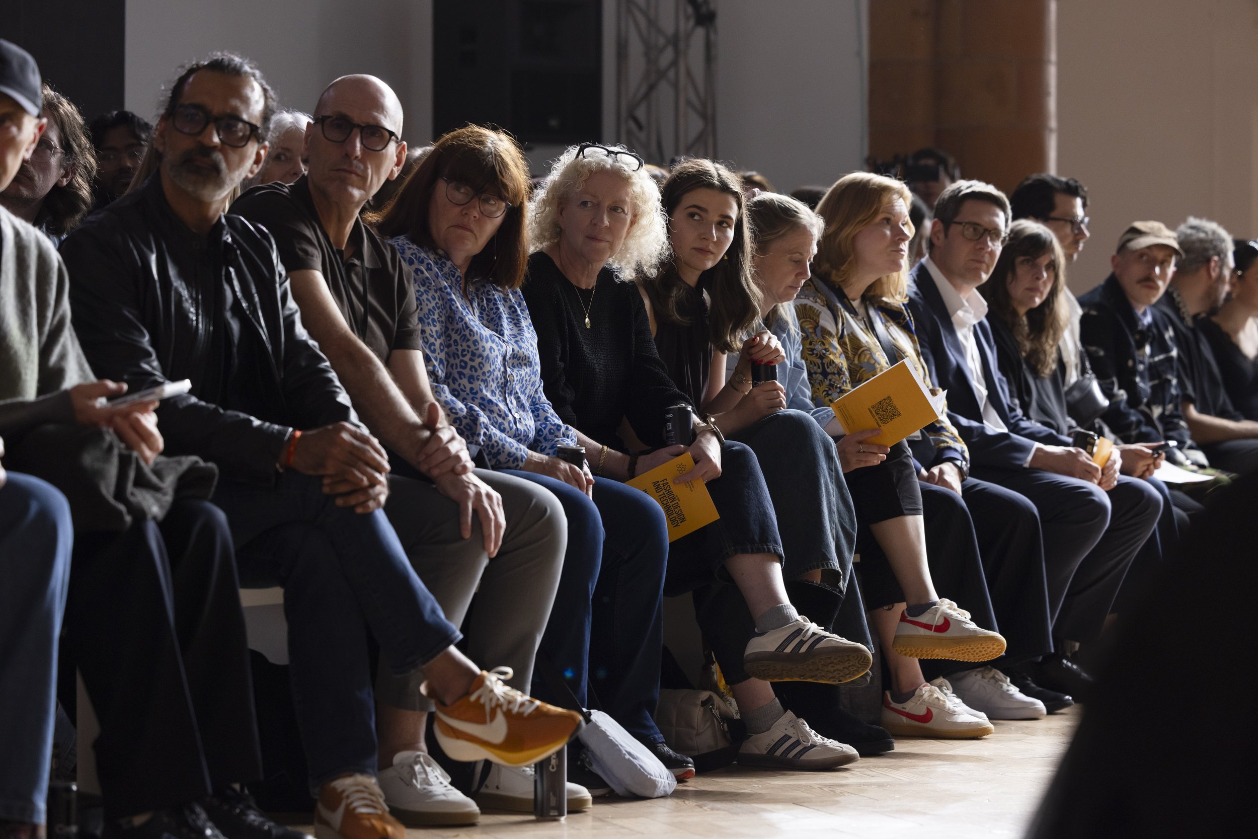 Backlit front-row audience seated at Manchester fashion show, silhouettes visible against runway lighting at Holden Gallery.