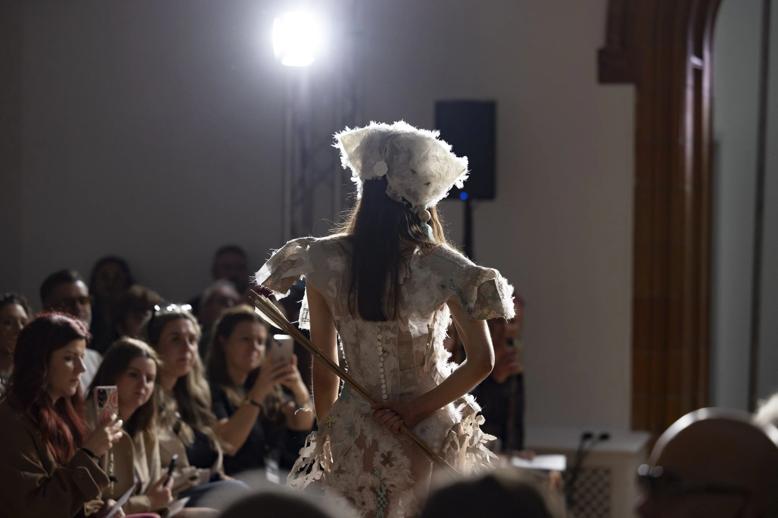 Rear view of a model walking down the runway at Manchester Fashion Institute INTERFACES showcase, holding two arrows behind her back as part of the presentation.