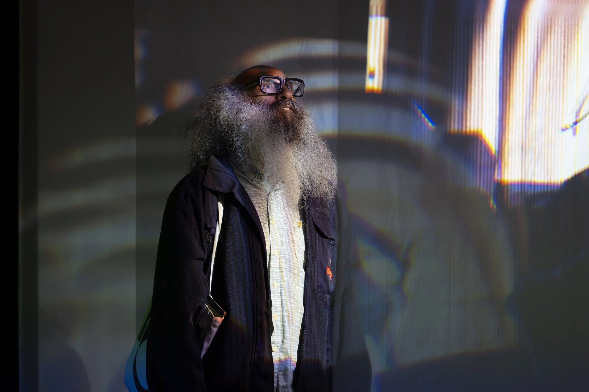 This image features a portrait of a man with a long, textured gray beard and glasses, standing before a light projection. He is wearing a dark jacket over a light-colored shirt and appears to be looking upward and to the side.