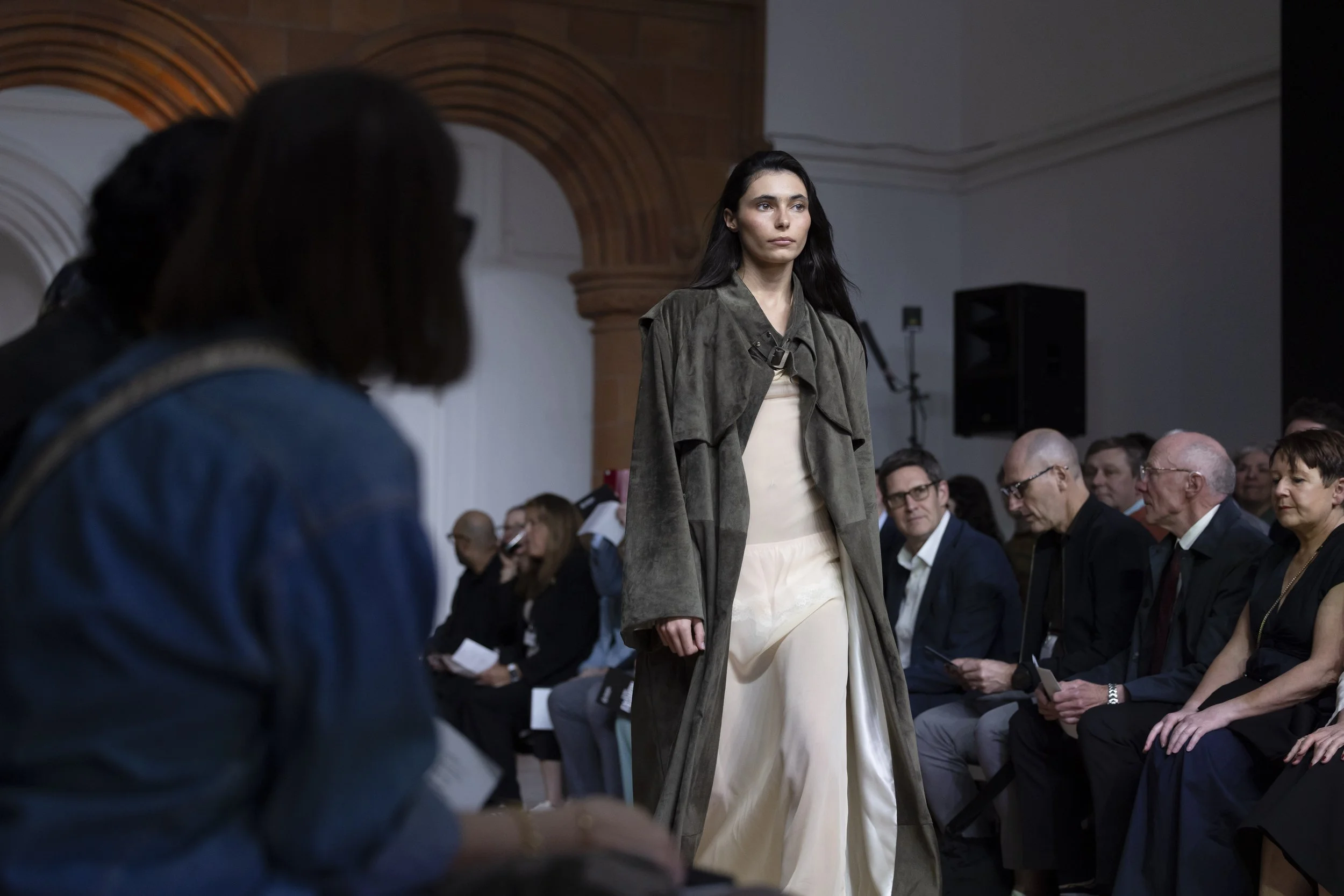 Model walking along the catwalk at MMU INTERFACES showcase, surrounded by audience seated on either side of the runway in a live fashion presentation.