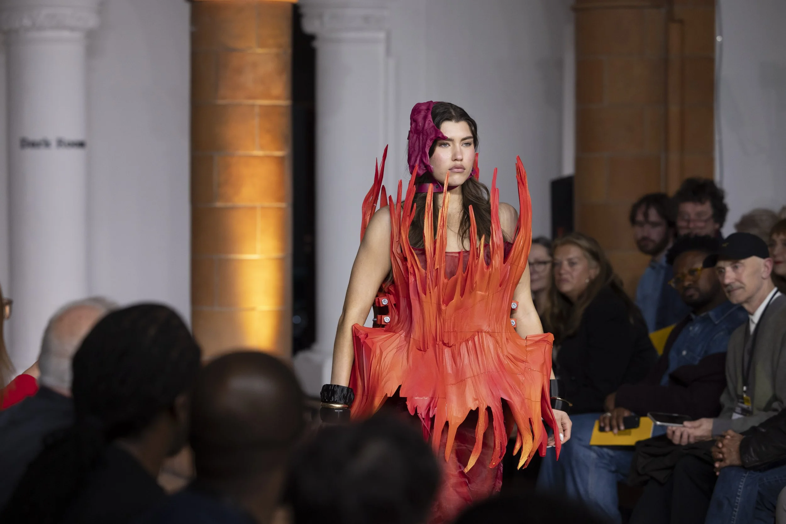 Model walking down the catwalk at Manchester Fashion Institute showcase in a flowing flaming orange dress, audience seated on either side of the runway.