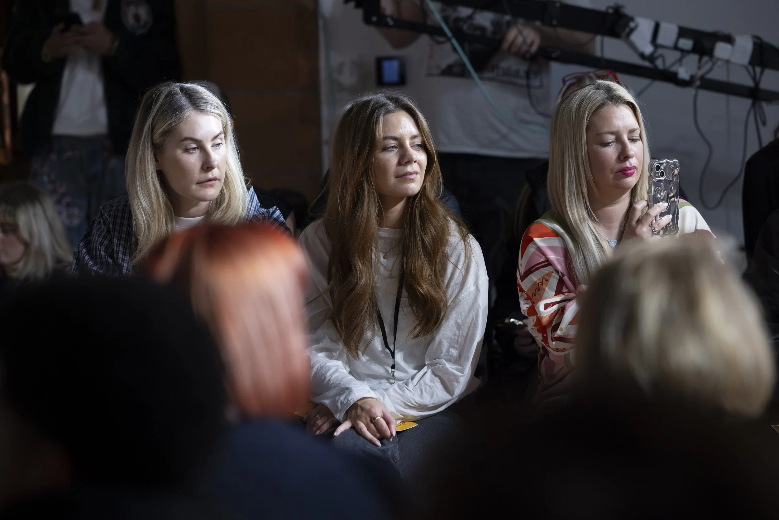 Three women seated in the audience at Manchester fashion showcase reviewing work presented during the runway event.