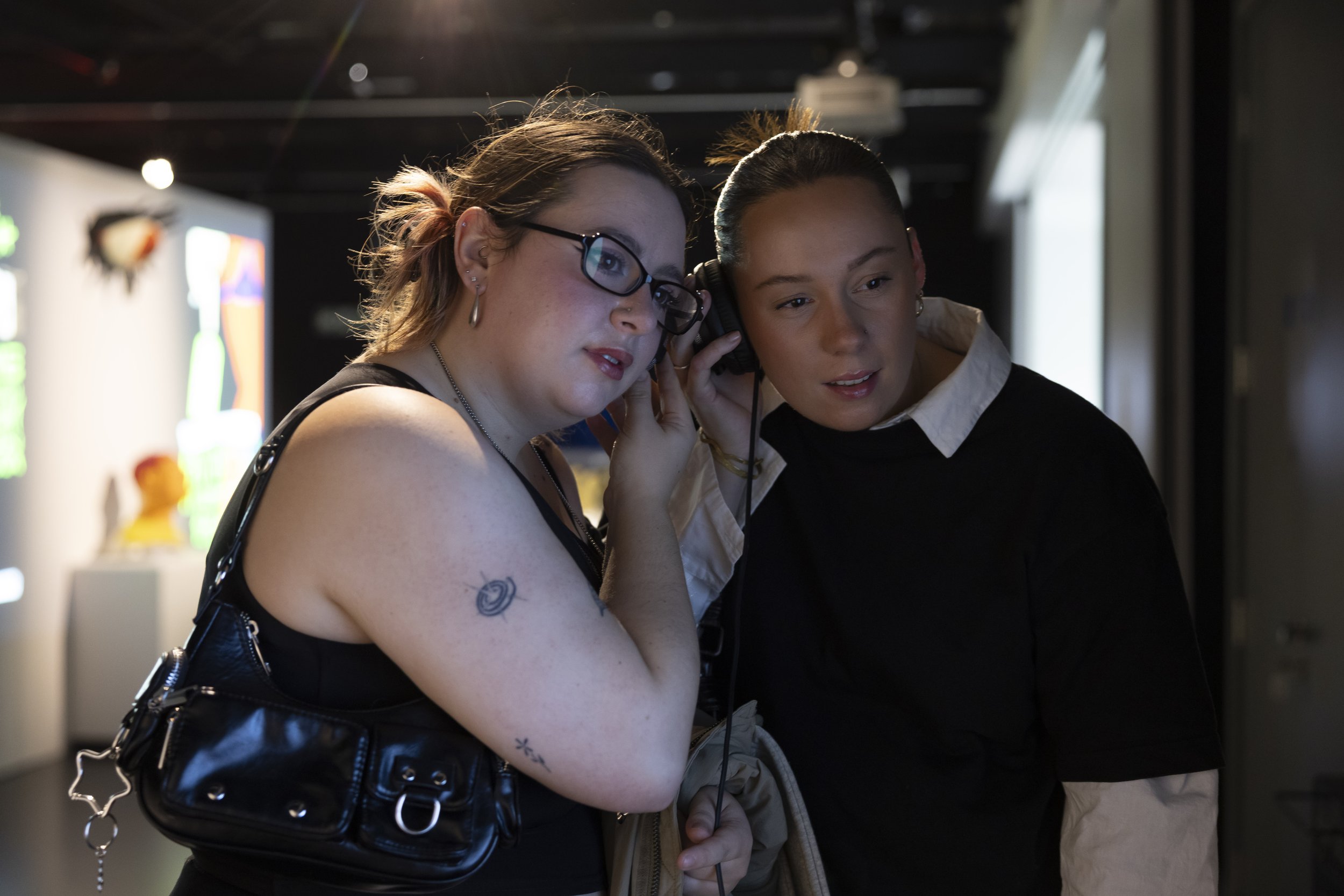 Two young women share headphones while watching a video artwork at Manchester Metropolitan University's More to Come exhibition, silhouetted against the projected image.