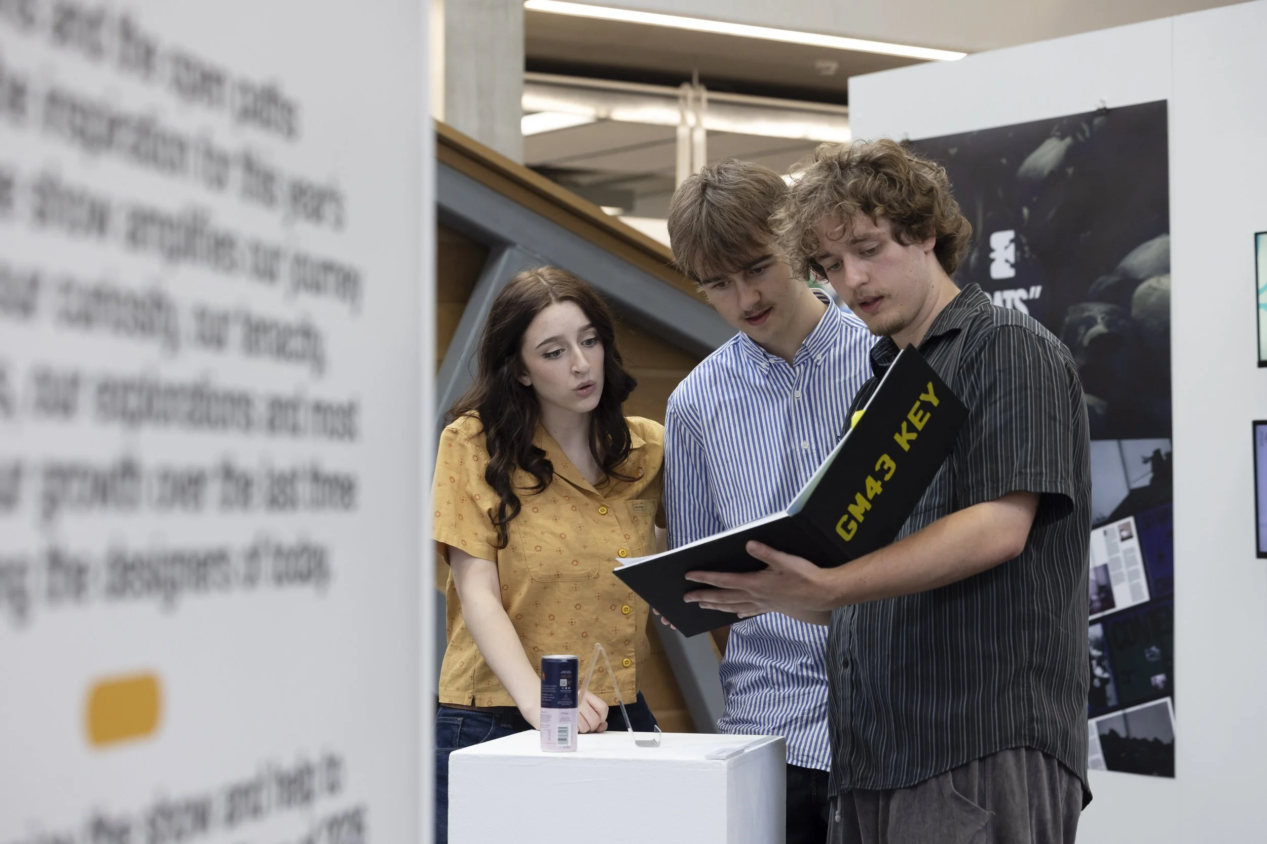 visitors during the exhibition's opening night private view at Manchester Metropolitan University's Oxford Road campus. One individual is holding a black portfolio or book with the text "GM43 KEY" displayed in yellow lettering on the cover.