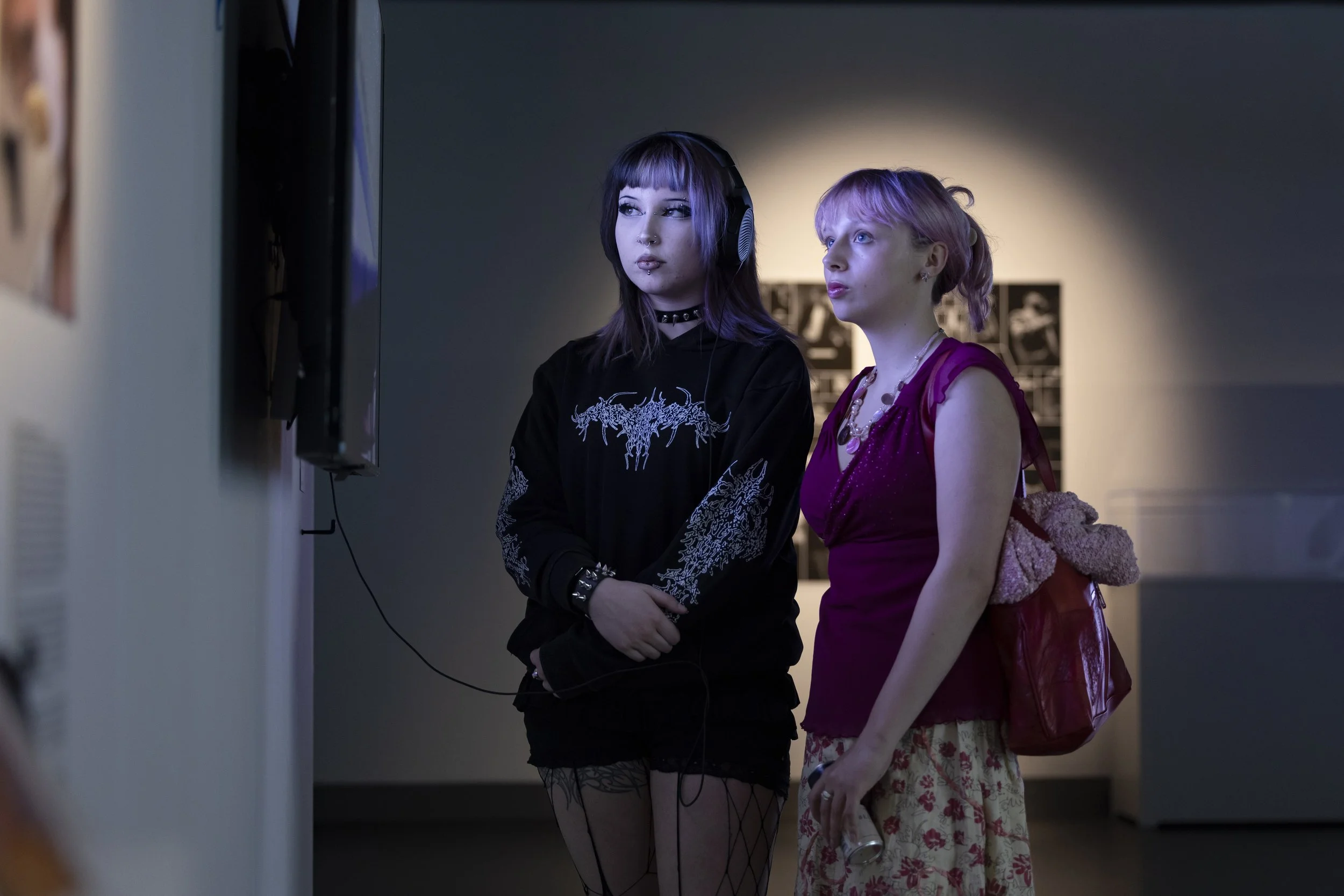Two young women with contrasting aesthetics—one in dark alternative fashion with a black graphic hoodie and fishnets, the other in a colorful bohemian-style pink top and floral skirt—stand together in an art gallery, focused on an exhibit.
