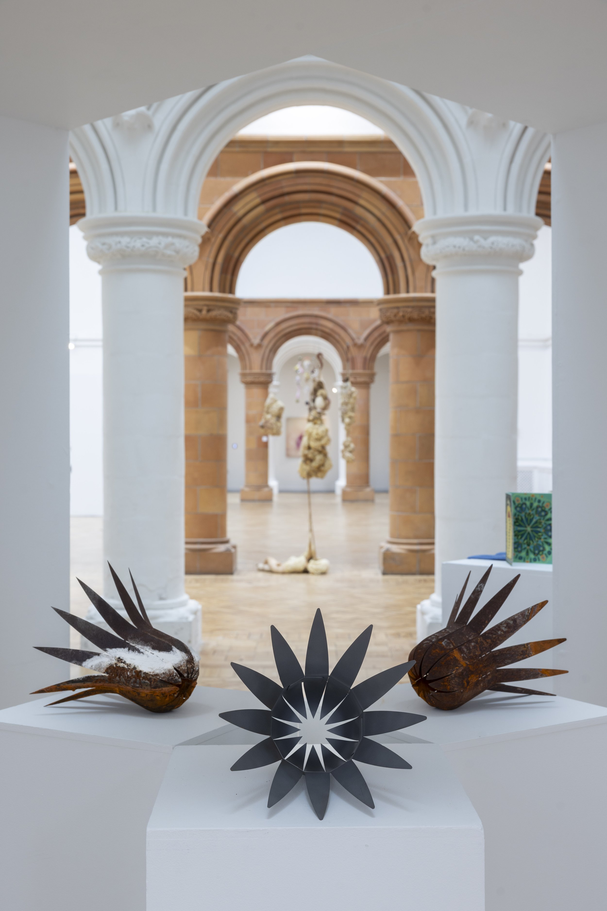 Three metallic sculptures displayed on a plinth, with other sculptural works in the background at the Holden Gallery postgraduate art exhibition.