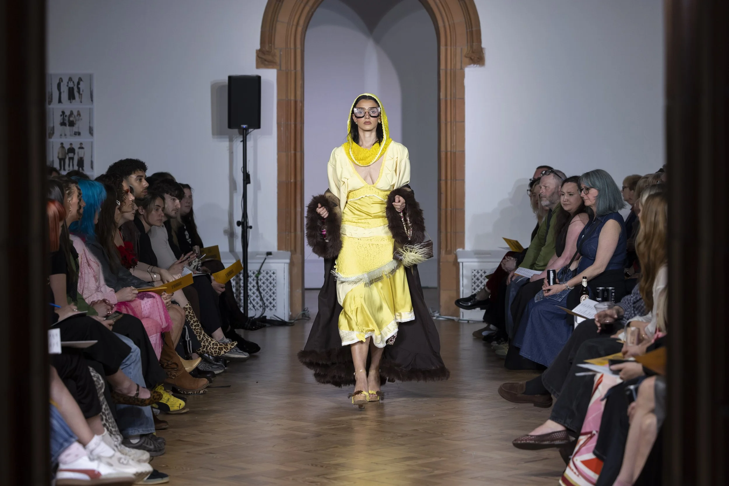 Fashion model in a yellow dress walking down the runway at MMU INTERFACES showcase, with audience watching from both sides of the catwalk.