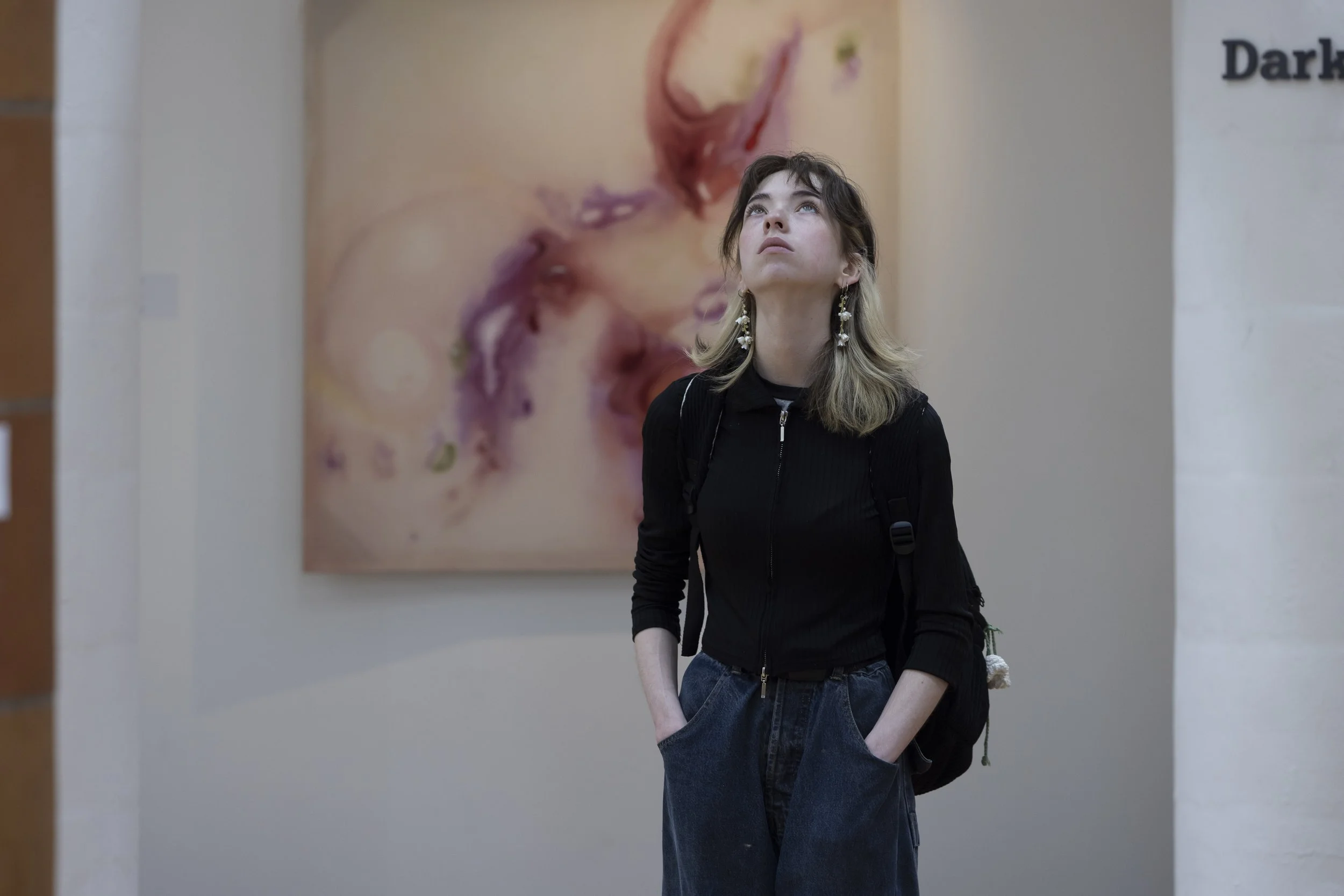Young woman looks up at suspended artwork hanging from the ceiling, with a pink painting in the background at the Holden Gallery, Manchester School of Art.