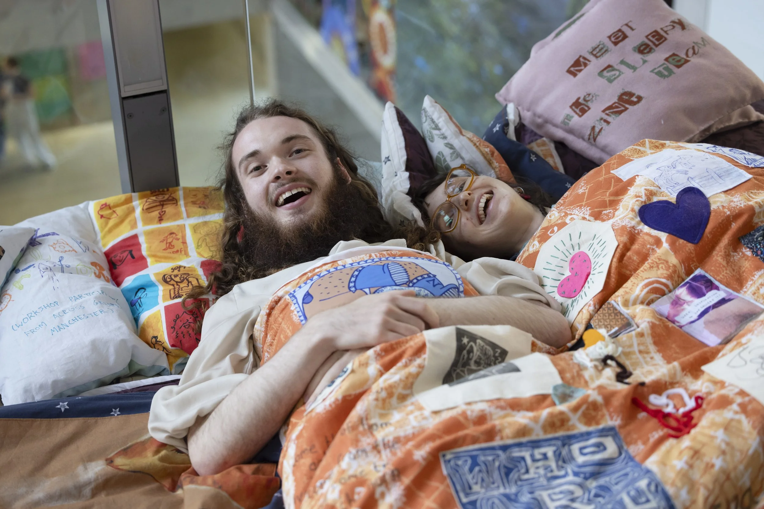 The image shows two people laughing while lying in an art installation that resembles a bed. The installation is part of the Manchester School of Art Degree Show 2026, specifically the opening night of an exhibition titled "More to Come".