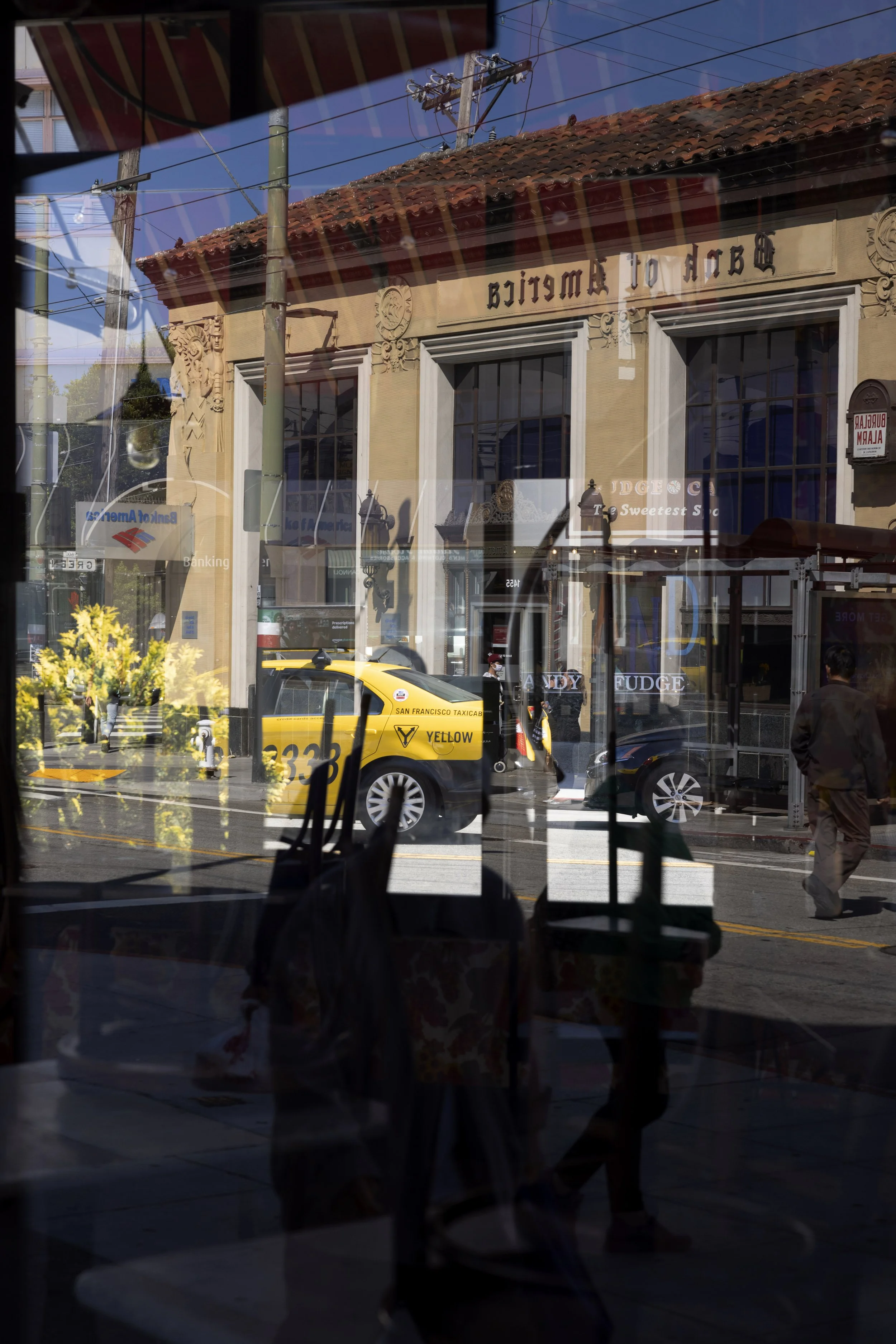 Abstract street scene in San Francisco seen through layered reflections in glass, showing fragments of Americana including a Bank of America sign, a yellow taxi, storefront candy displays, and passers-by in motion.