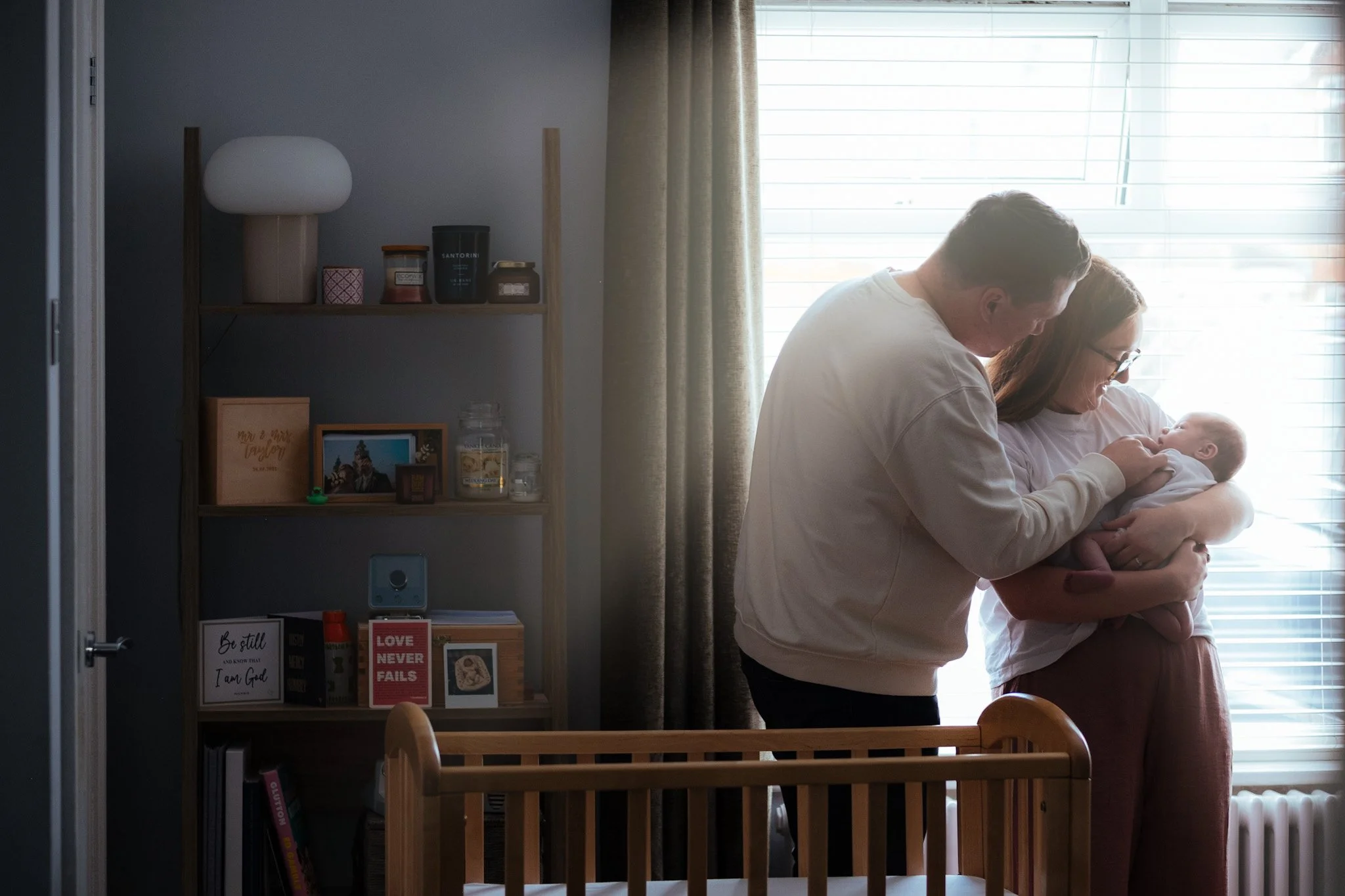 Intimate domestic scene — parent by nursery cot at window, family lifestyle photography