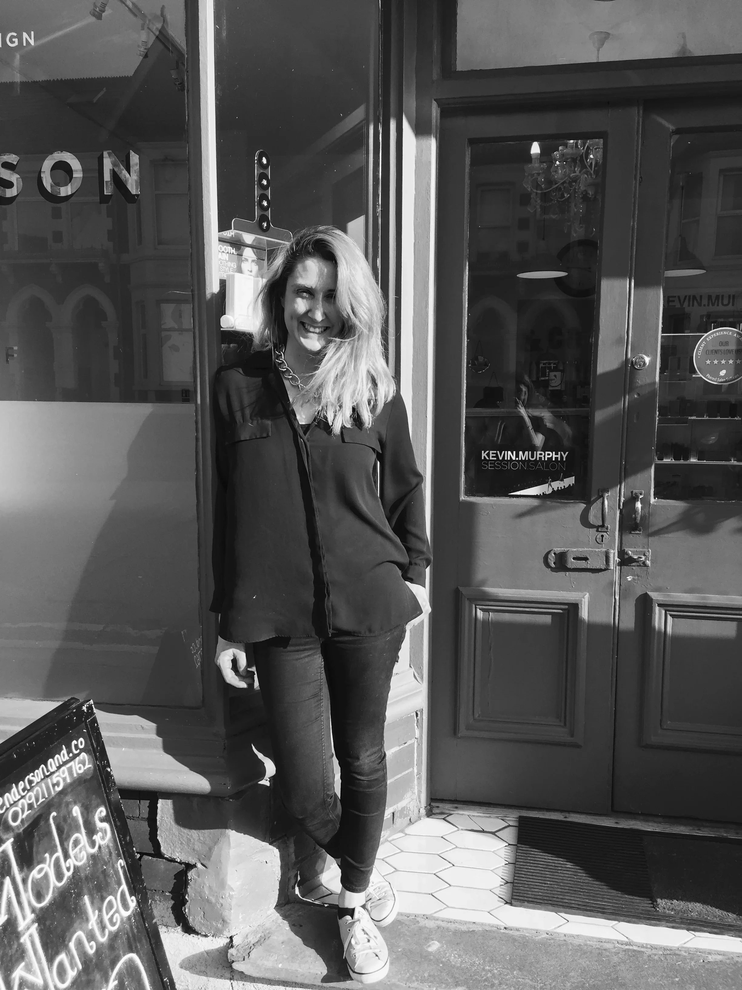 A woman with blonde hair, wearing a black shirt, jeans, and sneakers, standing outside a salon with a smile.