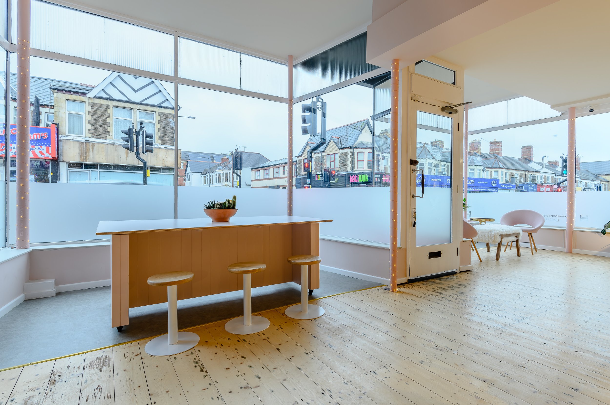 Empty cafe interior with wooden flooring, white walls, large windows, a small orange table with two bar stools, a white chair with a furry cushion, and a view of shops and houses outside.
