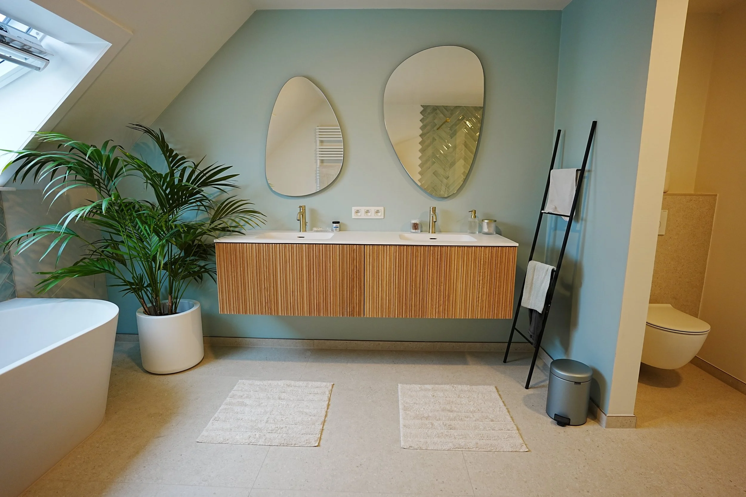 Moderne badkamer met een dubbele wastafel, twee spiegels, een grote plant in een witte pot, een vrijstaand bad, een ladderkast met handdoeken, een tandenborstel en een view op een toilet.