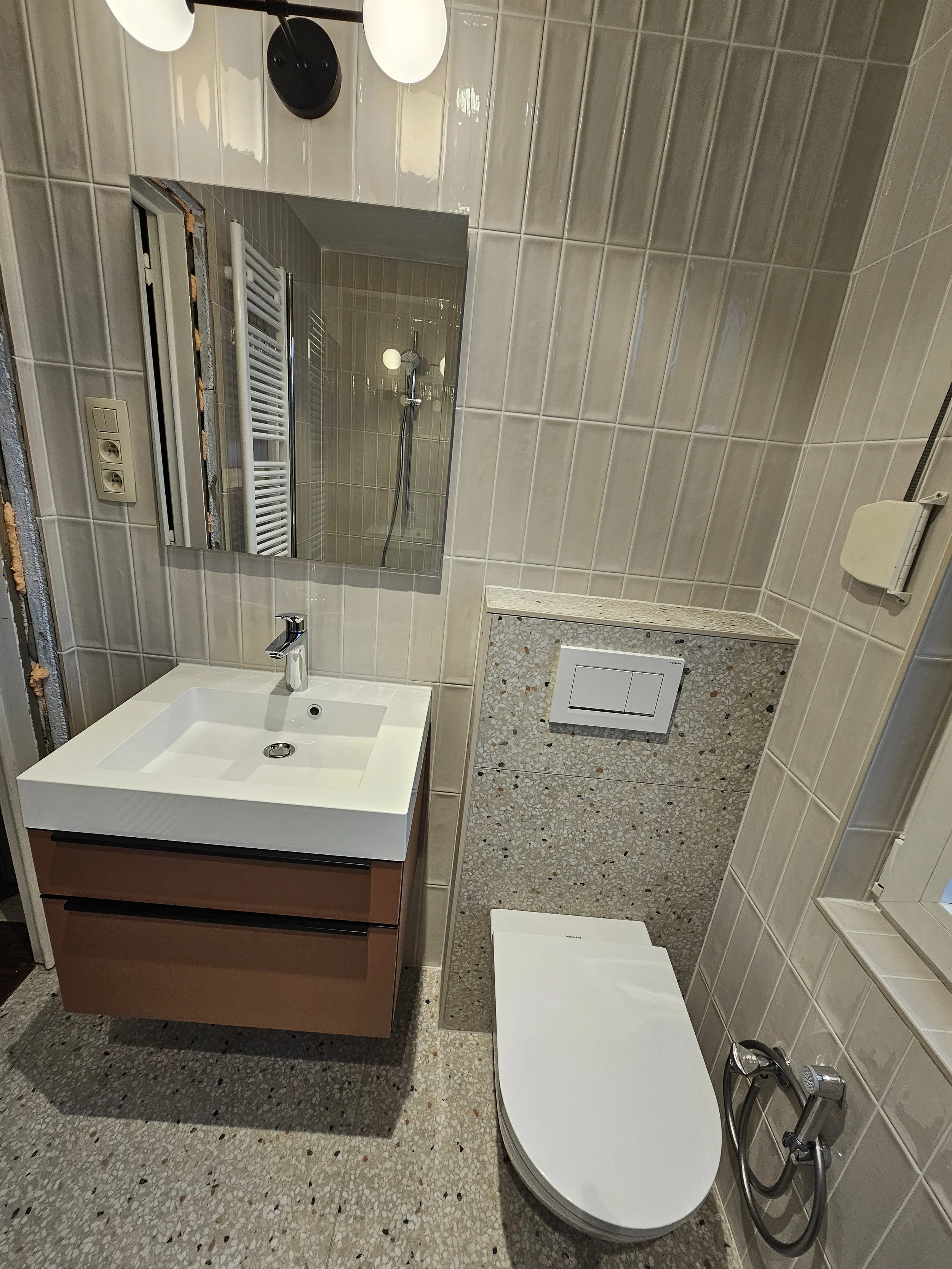 Een kleine badkamer met een spiegel, een wastafel met twee laden en een toilet. De muur is betegeld en er is een spiegelkast boven de wastafel. In de spiegel is ook een handdouche te zien.