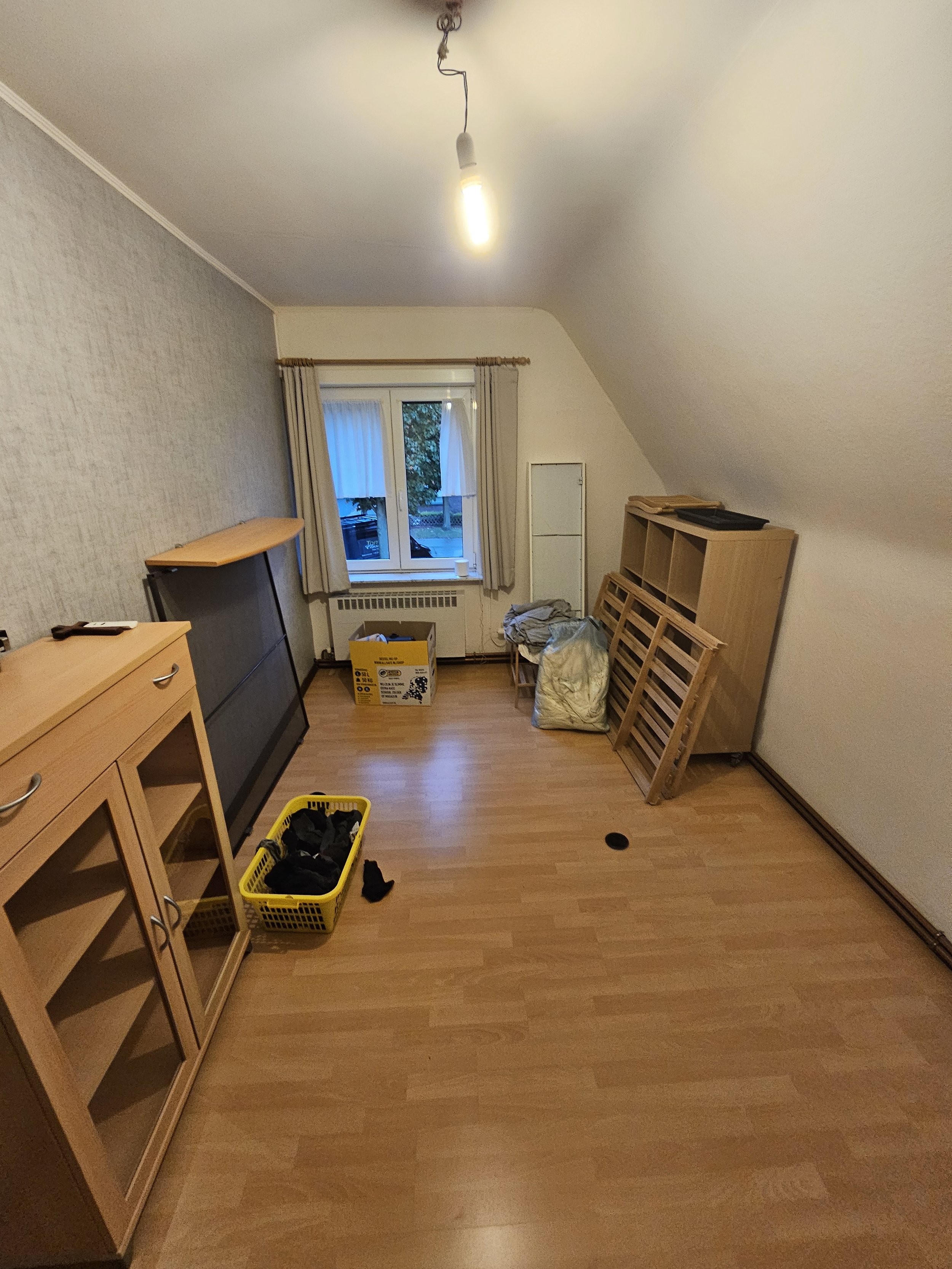 Leeg kamertje met houten meubels, dozen en spullen, raam met gordijnen, vloer van hout, plafond met blootliggende lamp, en een ventilatieschacht in de vloer.