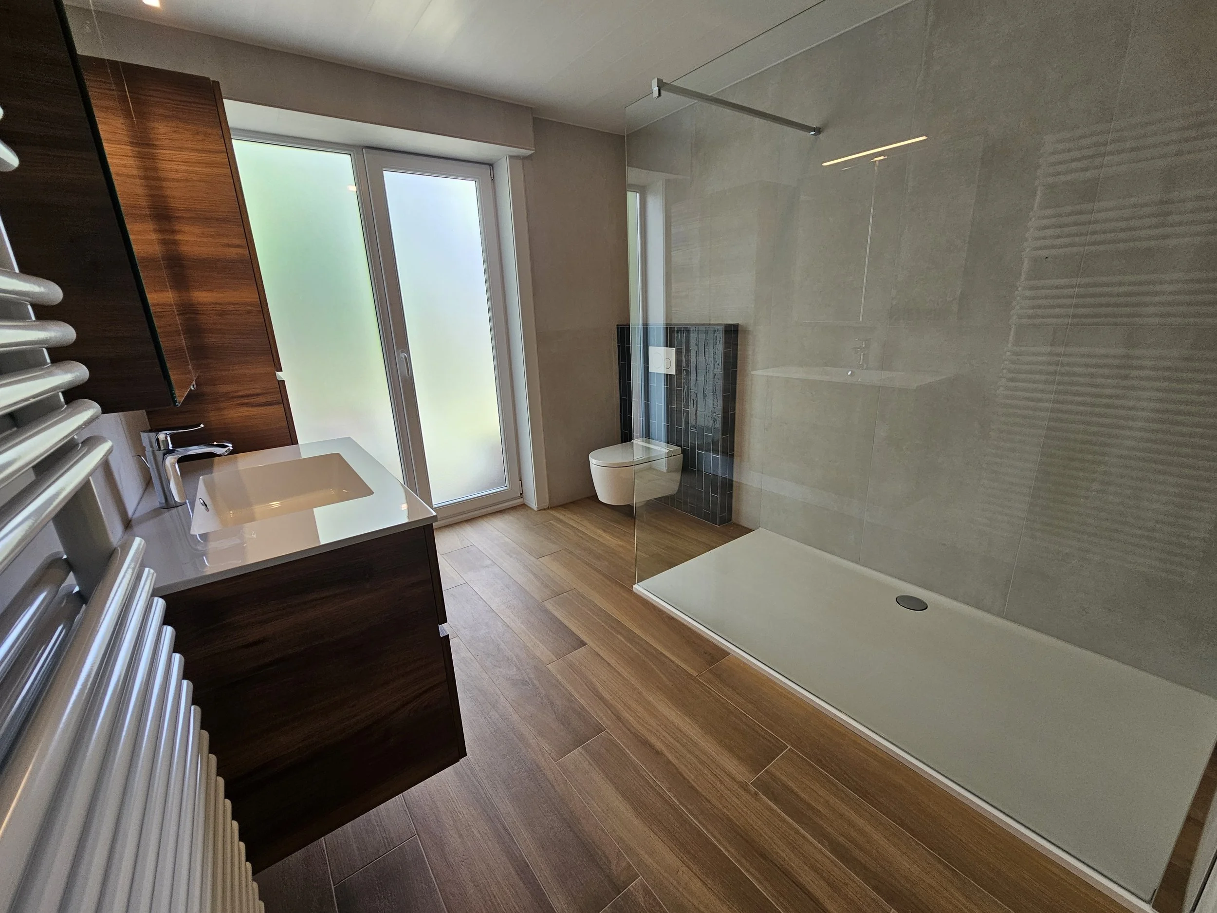 Een moderne badkamer met houten vloer, een grote inloopdouche met glazen wand, een muurhangend toilet en een fontein met een witte keramieken wastafel, naast een raam met frosted glas.