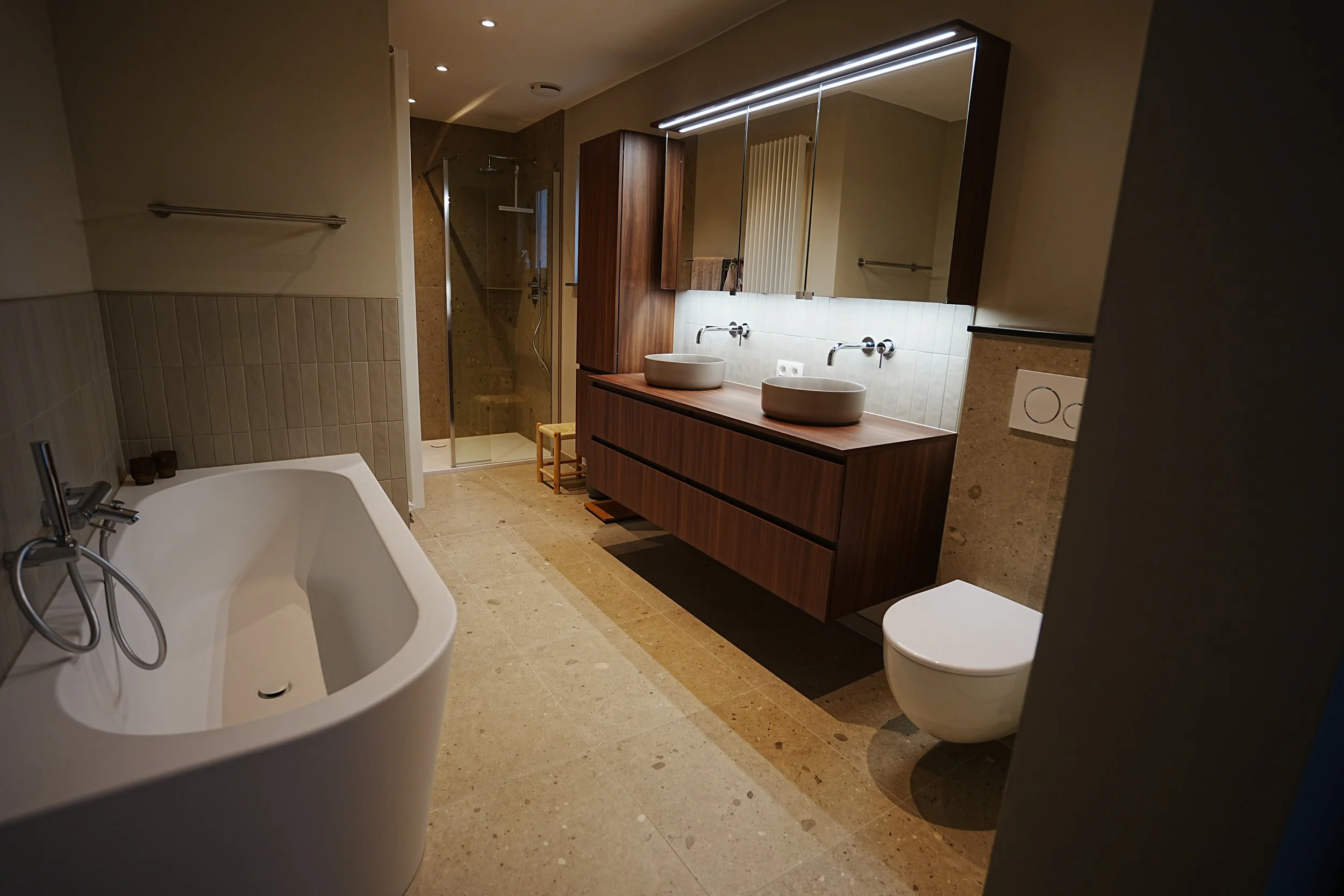 Moderne badkamer met een vrijstaand wit bad, dubbele wastafels in een houten kast, een toilet, en een inloopdouche met glazen deuren.