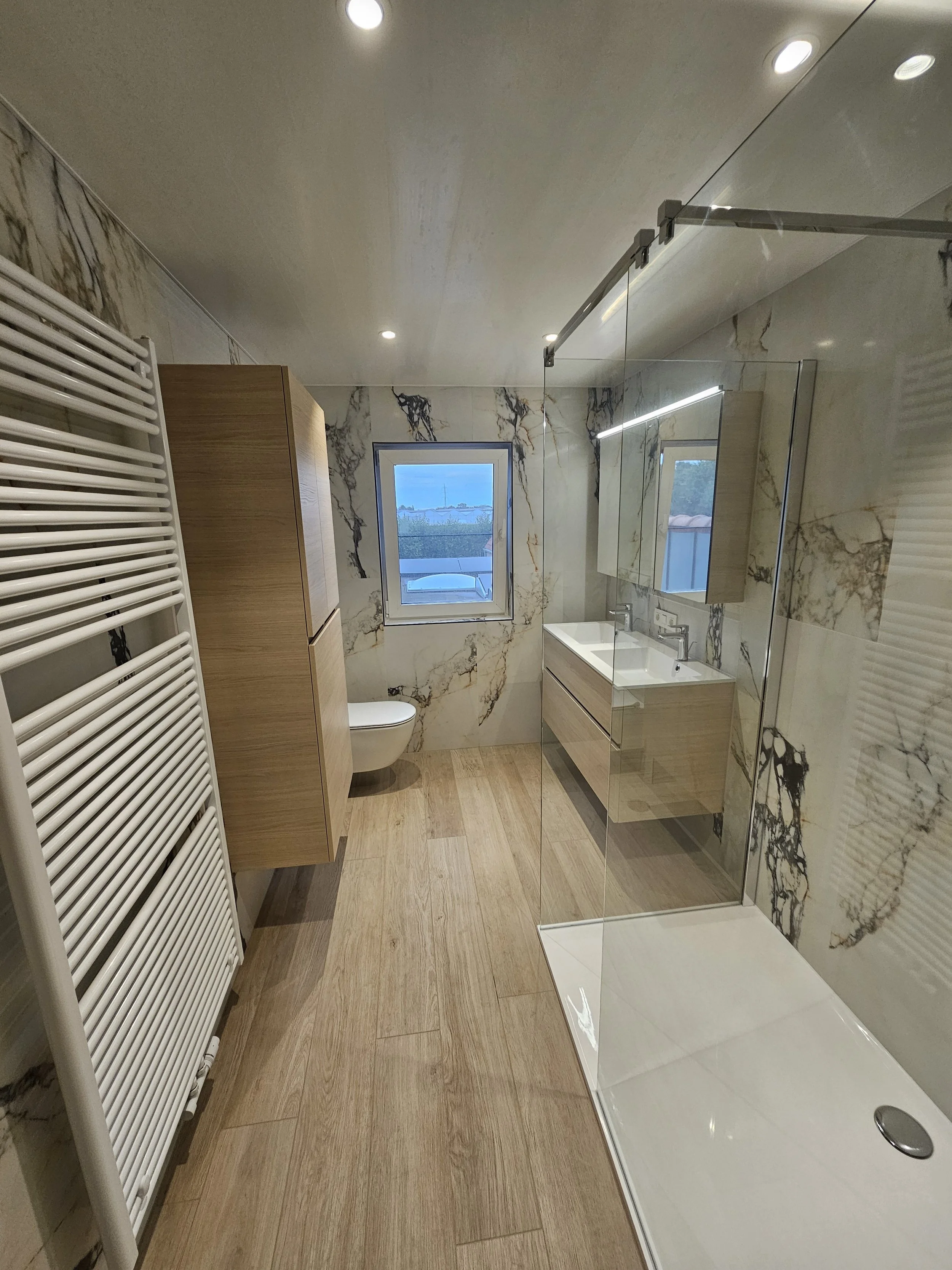 Een badkamer met een houten vloer, een witte radiator, een toilet, een raam, een houten wastafel met spiegel, en een inloopdouche met glazen wand.