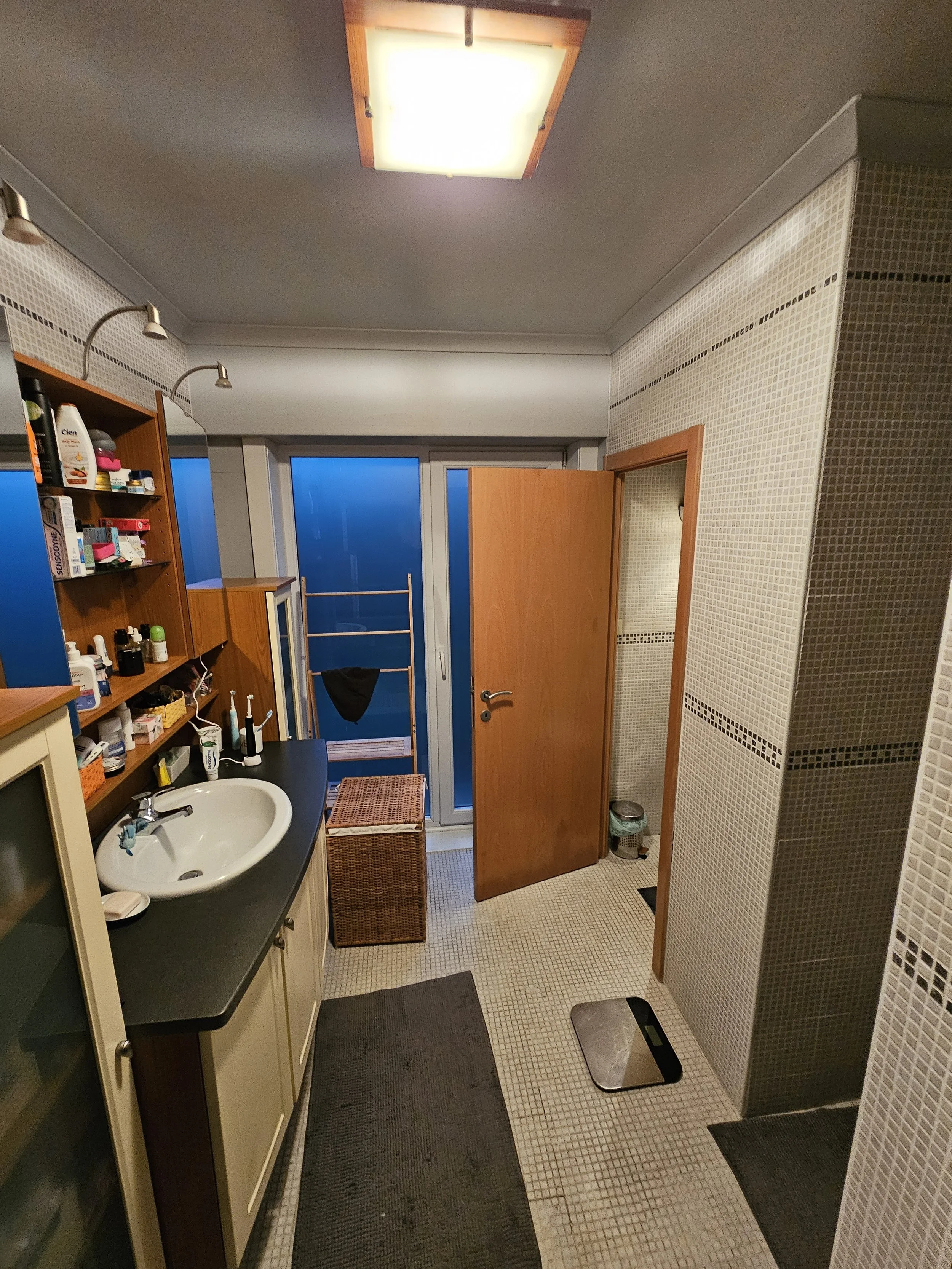 Binnenkant van een badkamer met een wastafel, wandplanken met toiletartikelen, een verwarming, een open deur, een digitale weegschaal op de vloer, en een wasmand.