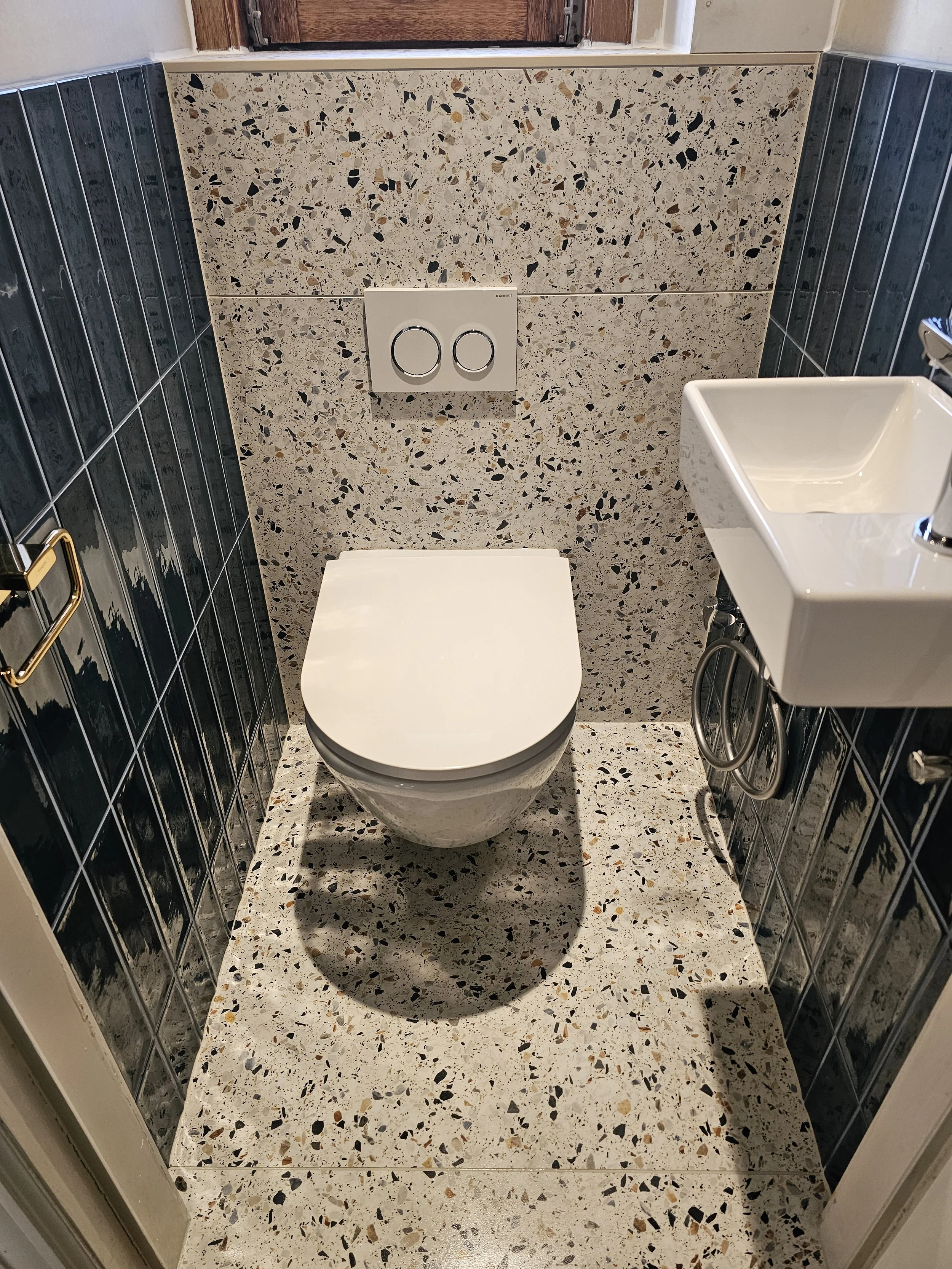 Klein toilet met muur en vloer bedekt met terrazzo tegels, een inbouwspüler, en een kleine wasbak aan de rechterkant.