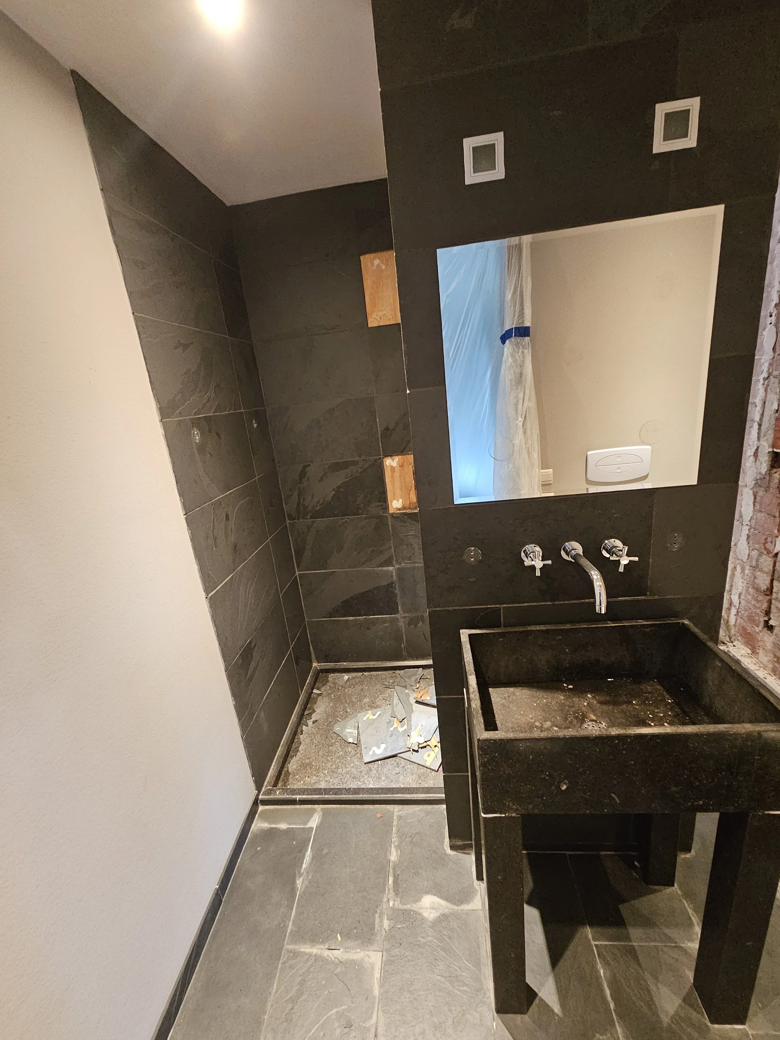 Een badkamer die wordt ingericht met zwarte tegels, een zwart wasbakje, een spiegel en een raam met plastic afdekking. Onderaan ligt vuil en bouwmateriaal.